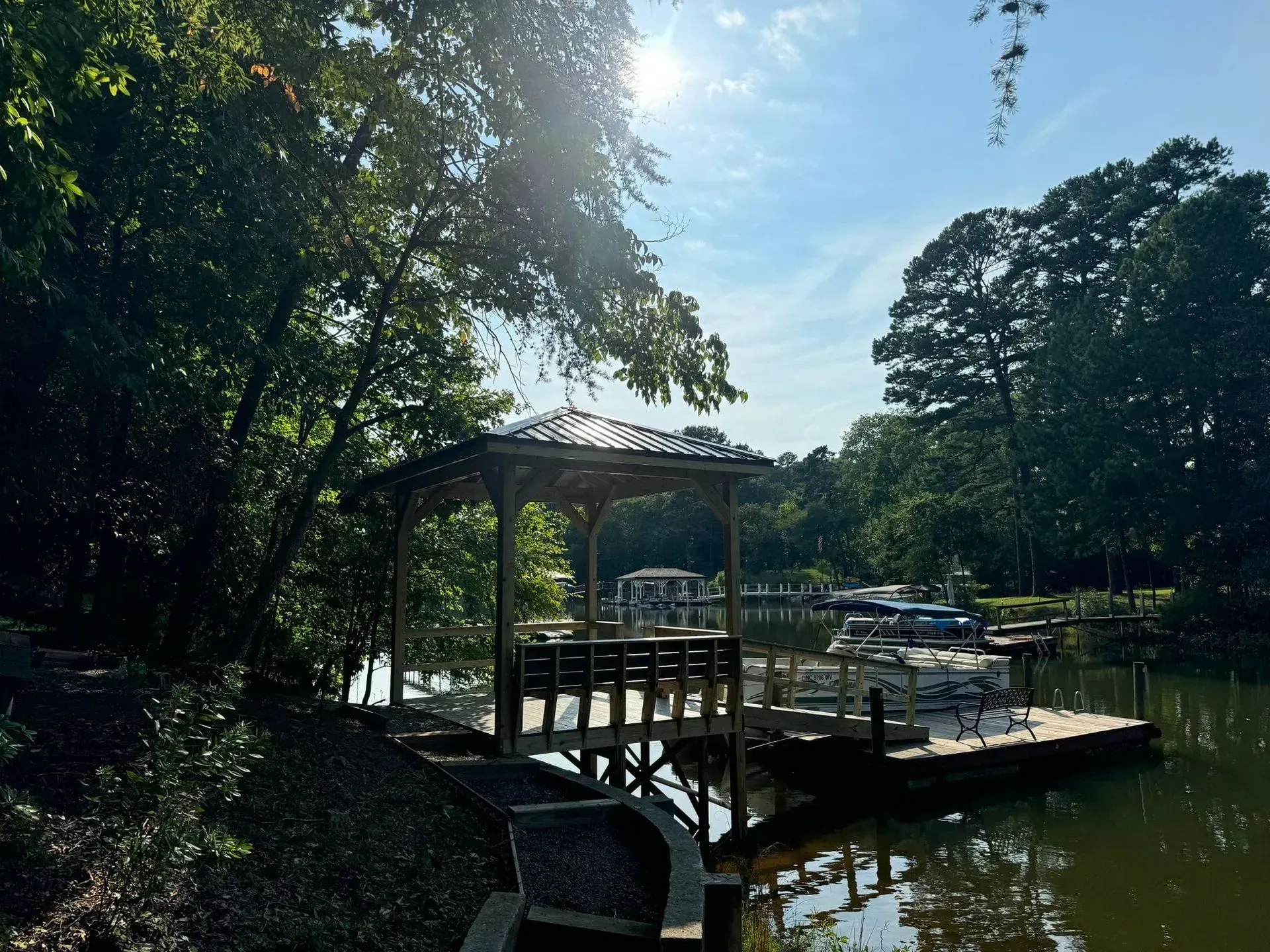 Dock with gazebo on a lake, surrounded by trees, bright sun in the sky.