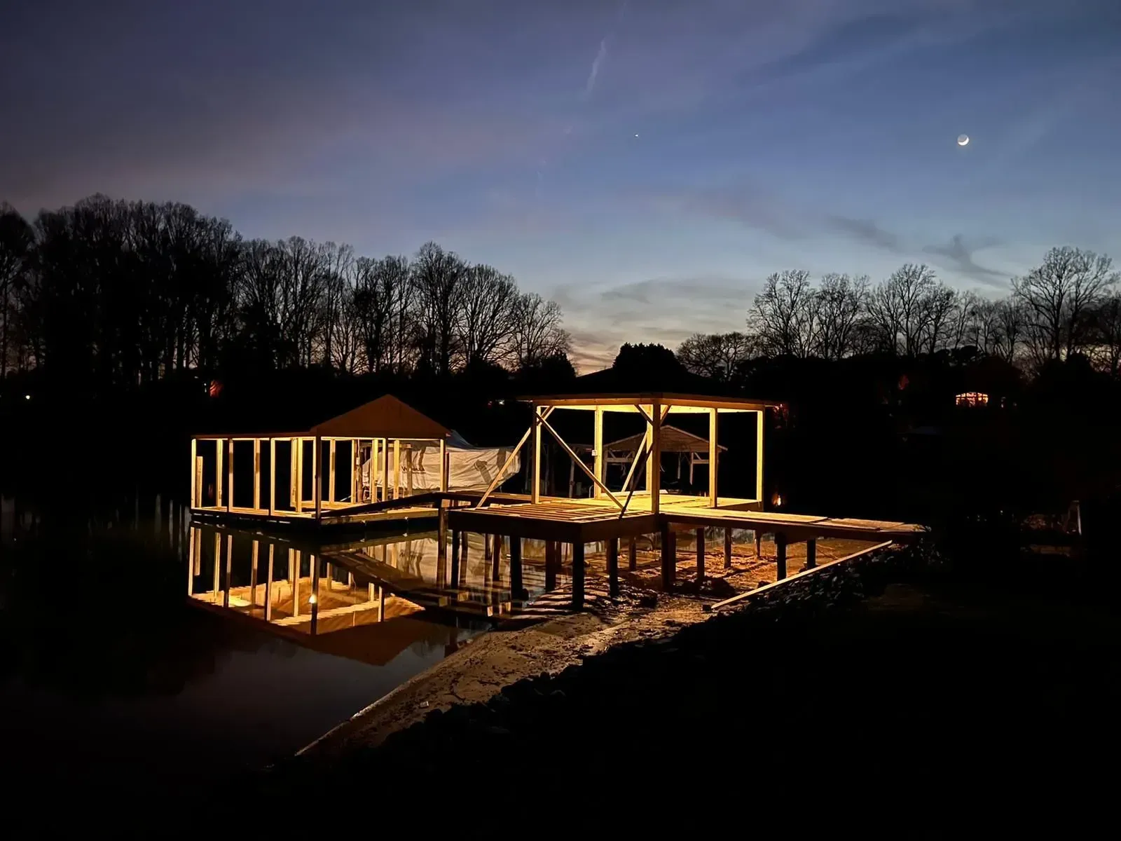 Lit wooden dock with gazebos reflecting in dark water at dusk. Trees and twilight sky in background.