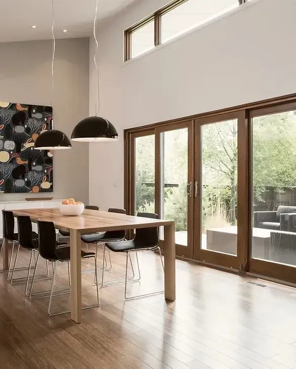 Modern dining room with wood table, black chairs, and large windows overlooking a backyard.