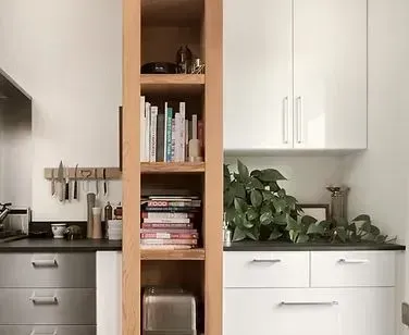 Kitchen with built-in wooden bookshelf between white cabinets. Books and a toaster on shelves, plant on countertop.