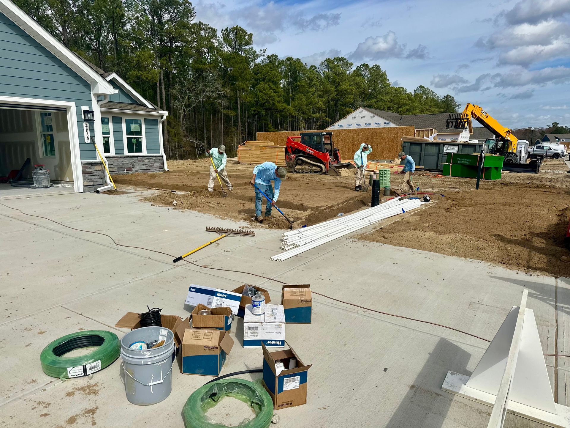 Construction site with workers digging, grading, and installing pipes near new houses under a cloudy sky.