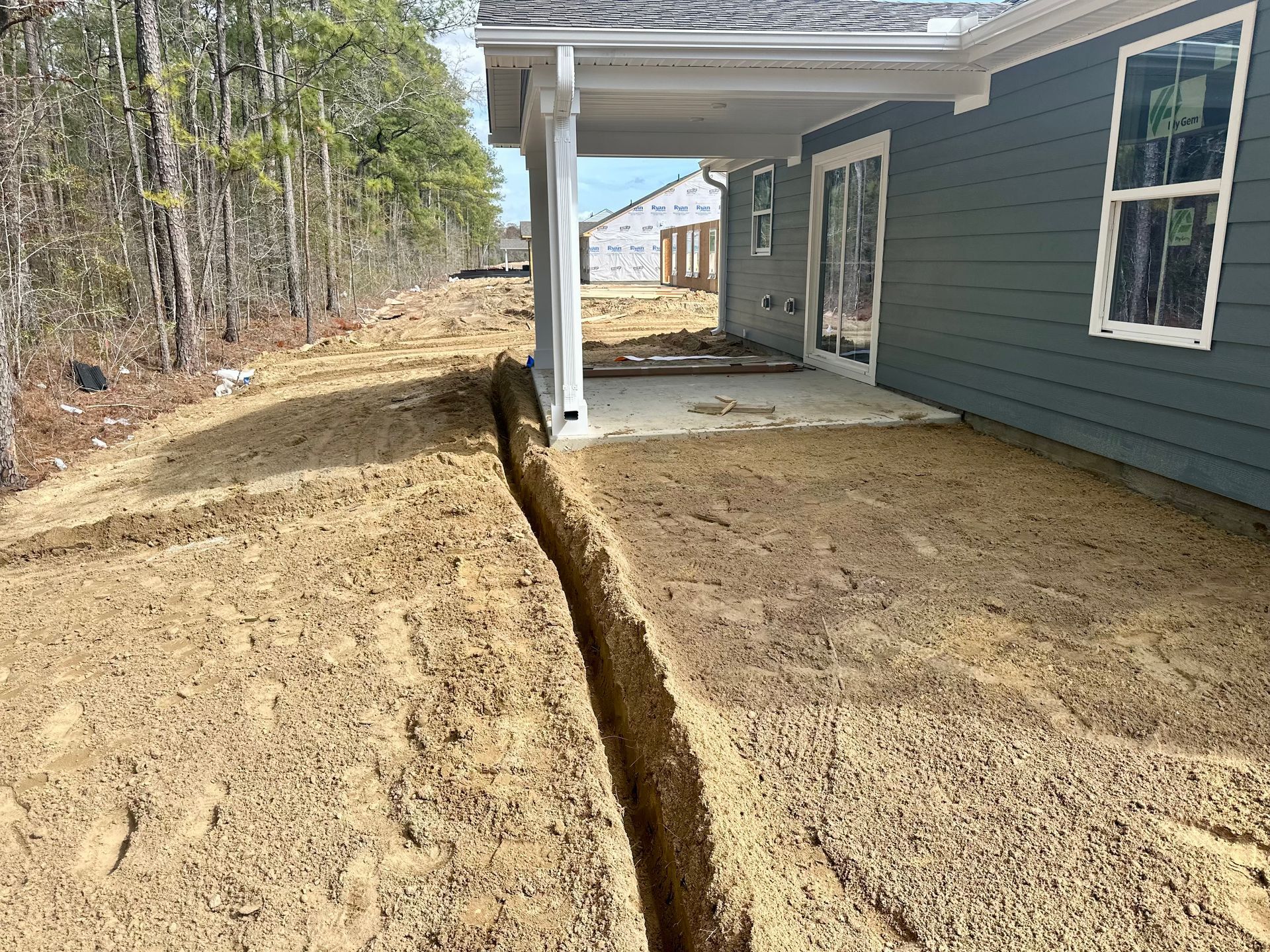 Trench dug near a house with a covered patio. The house is painted blue with a white window.