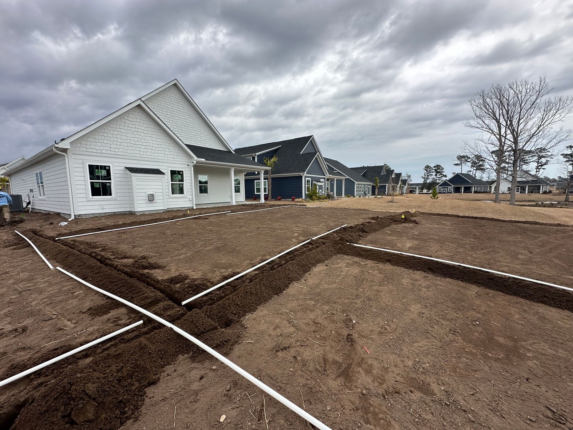 Trenches with white pipes in front of new homes under cloudy sky.