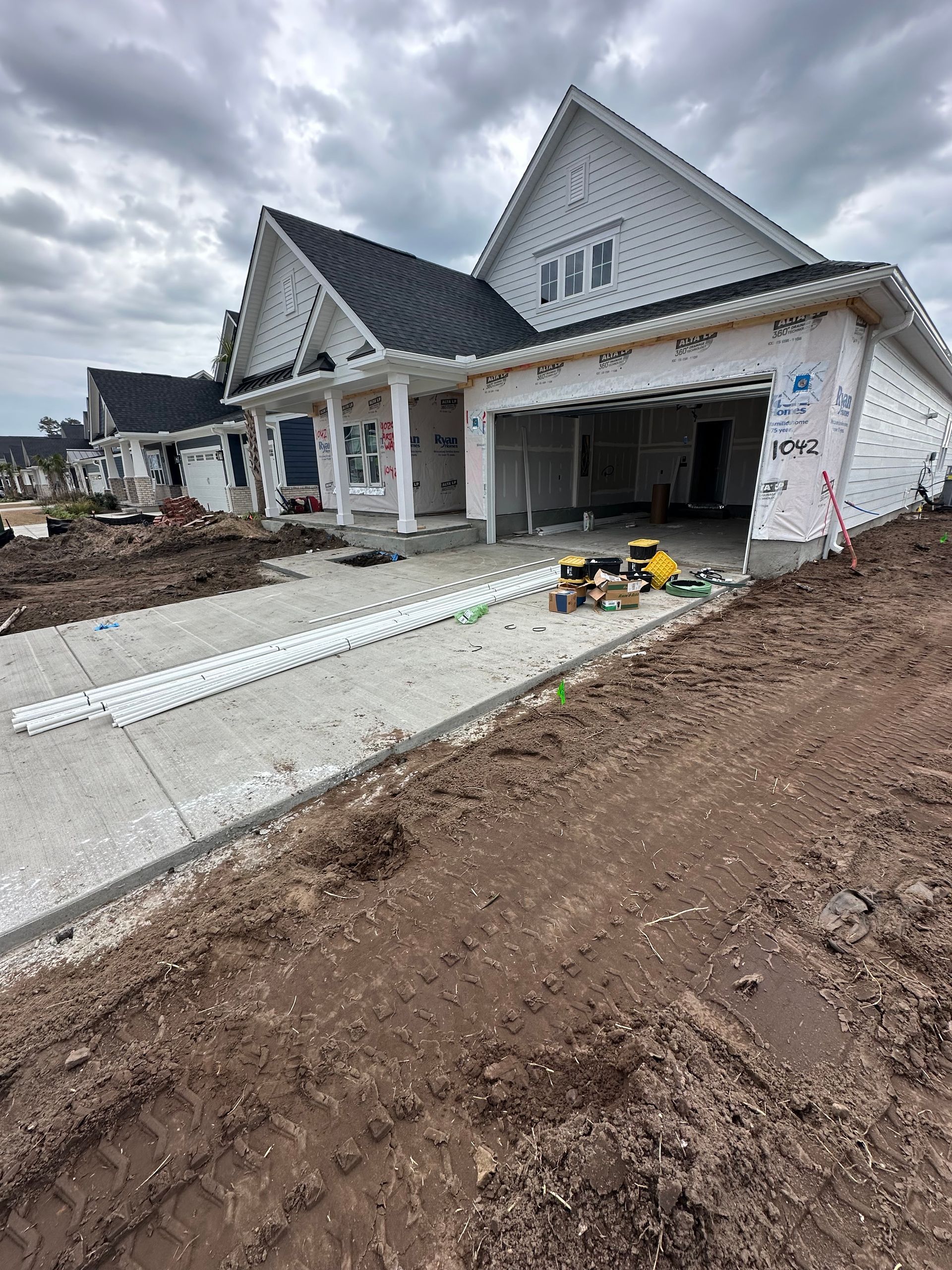 New house under construction with garage open, driveway, and brown soil. White siding, black roof.