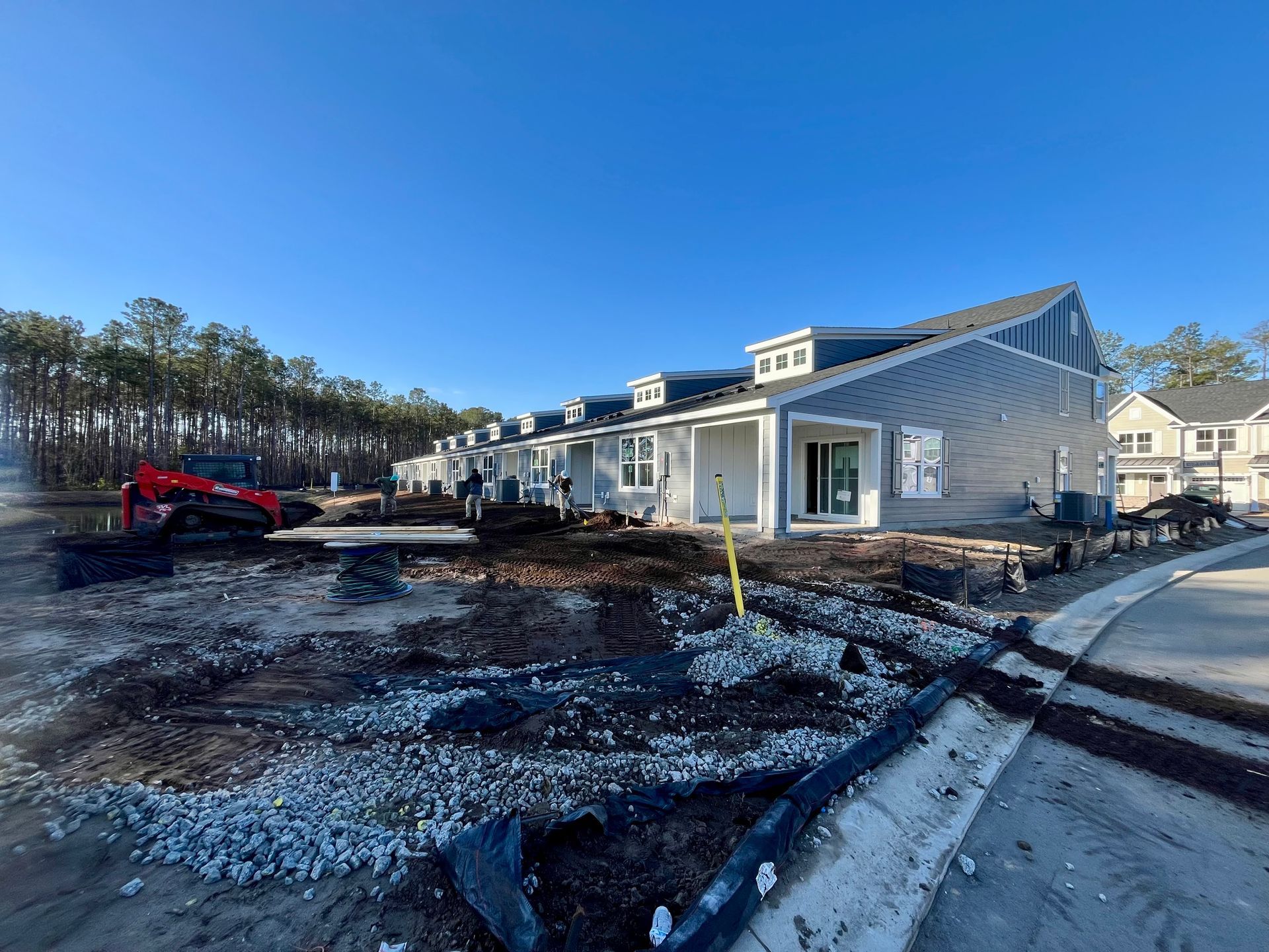 New construction townhomes on a sunny day. Dirt and gravel in the foreground; a red excavator is visible.
