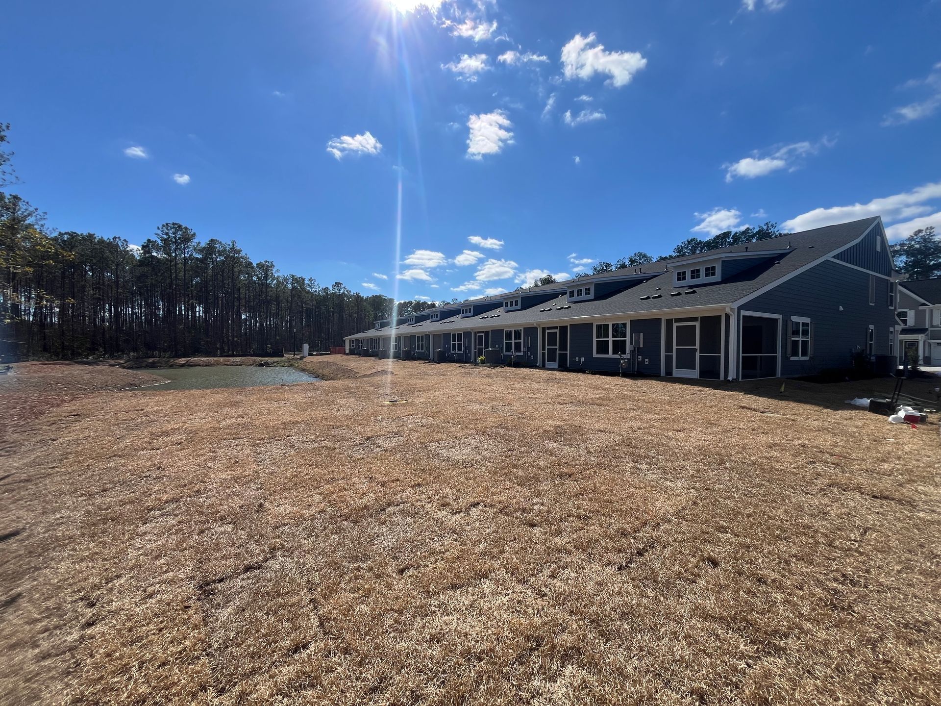 Blue apartment buildings with dark roof, surrounded by wood chips and trees under a sunny sky.