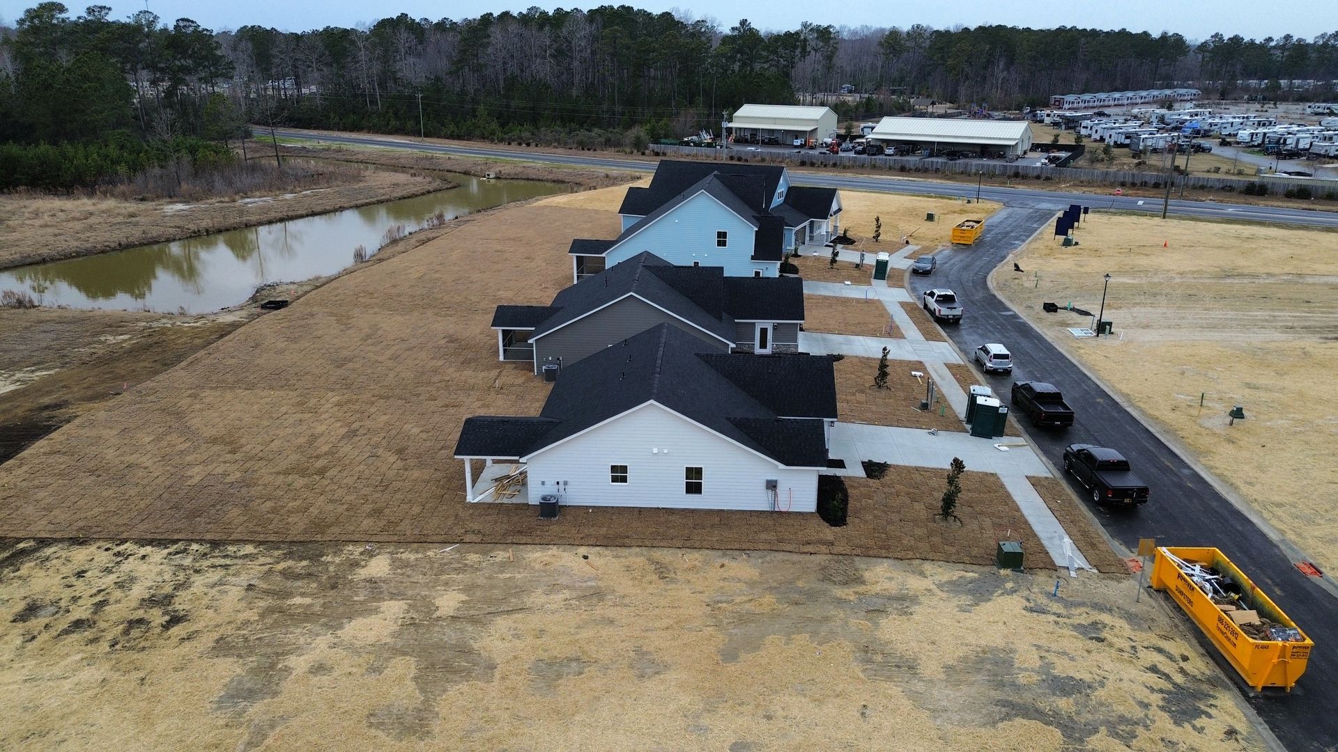 Row of new houses with dark roofs and light siding under construction, with a road and surrounding brown fields.
