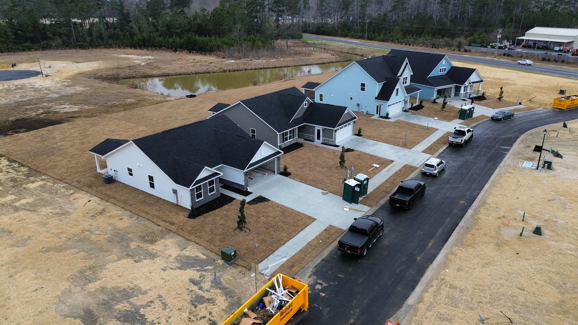 New homes under construction, brown and blue exteriors, vehicles on the street, and a pond.
