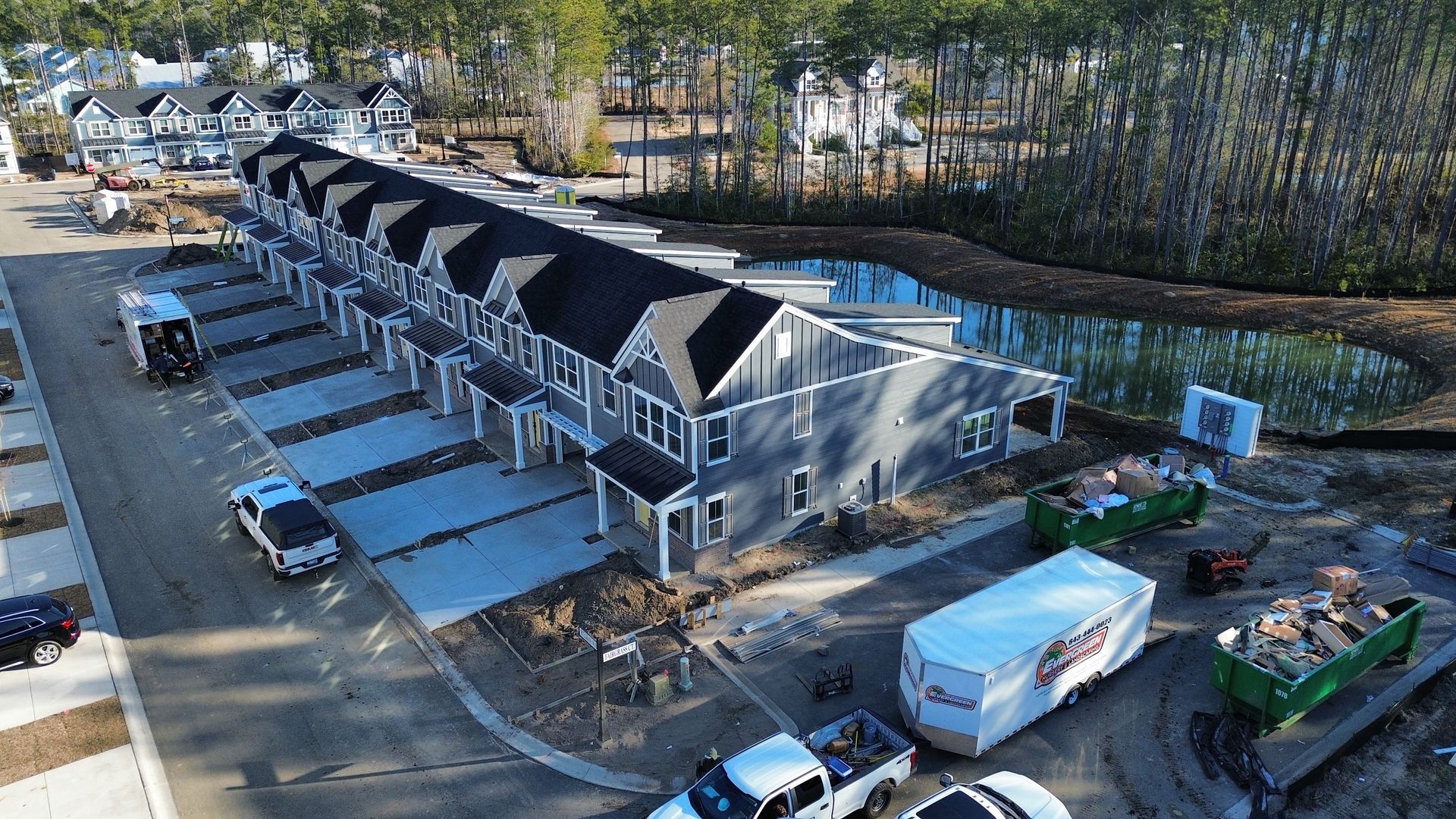 New townhouses under construction, gray siding, black roofs, and a small pond, surrounded by trees.