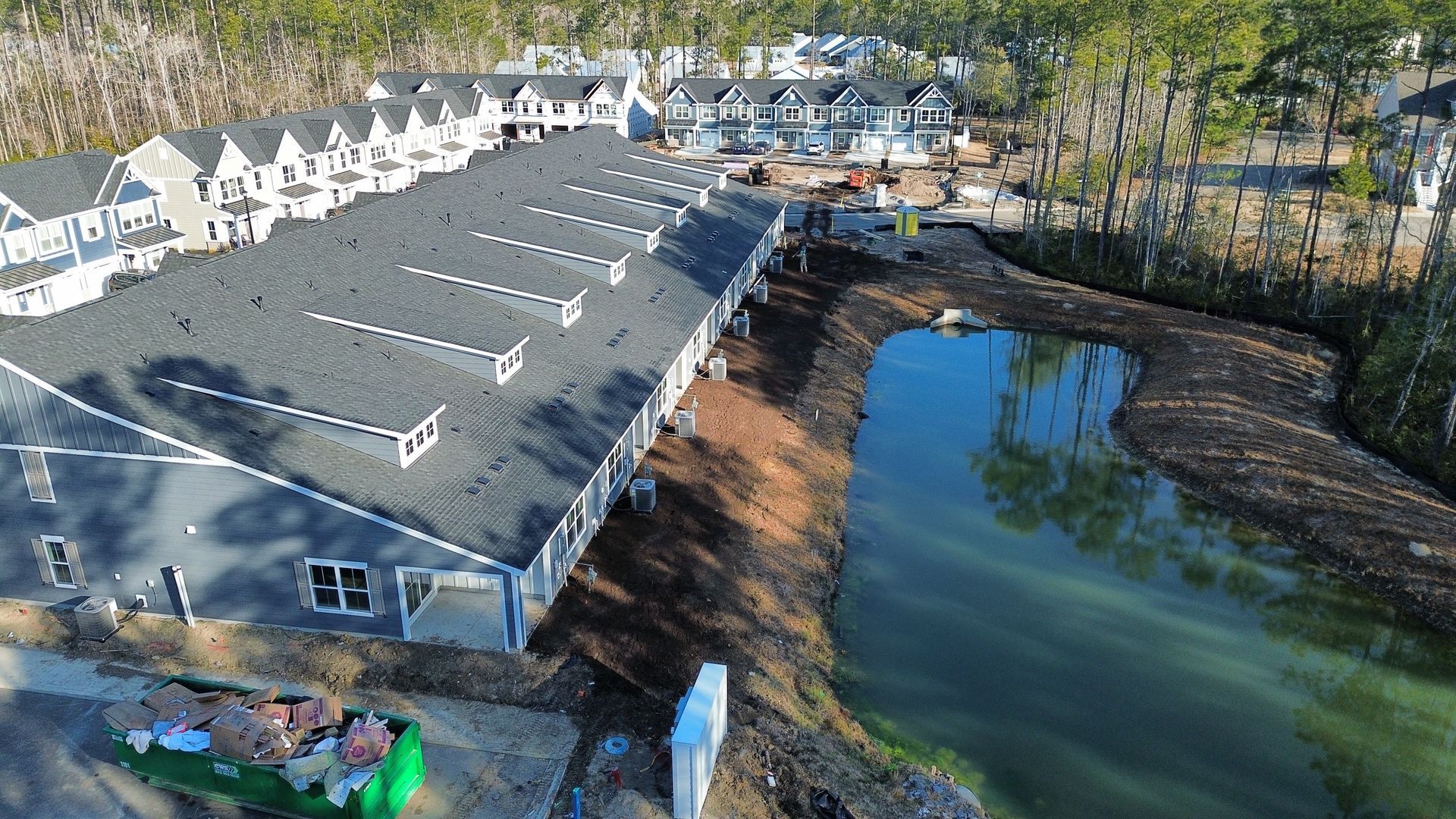 Aerial view of newly constructed townhouses with dark gray roofs, near a pond and trees.