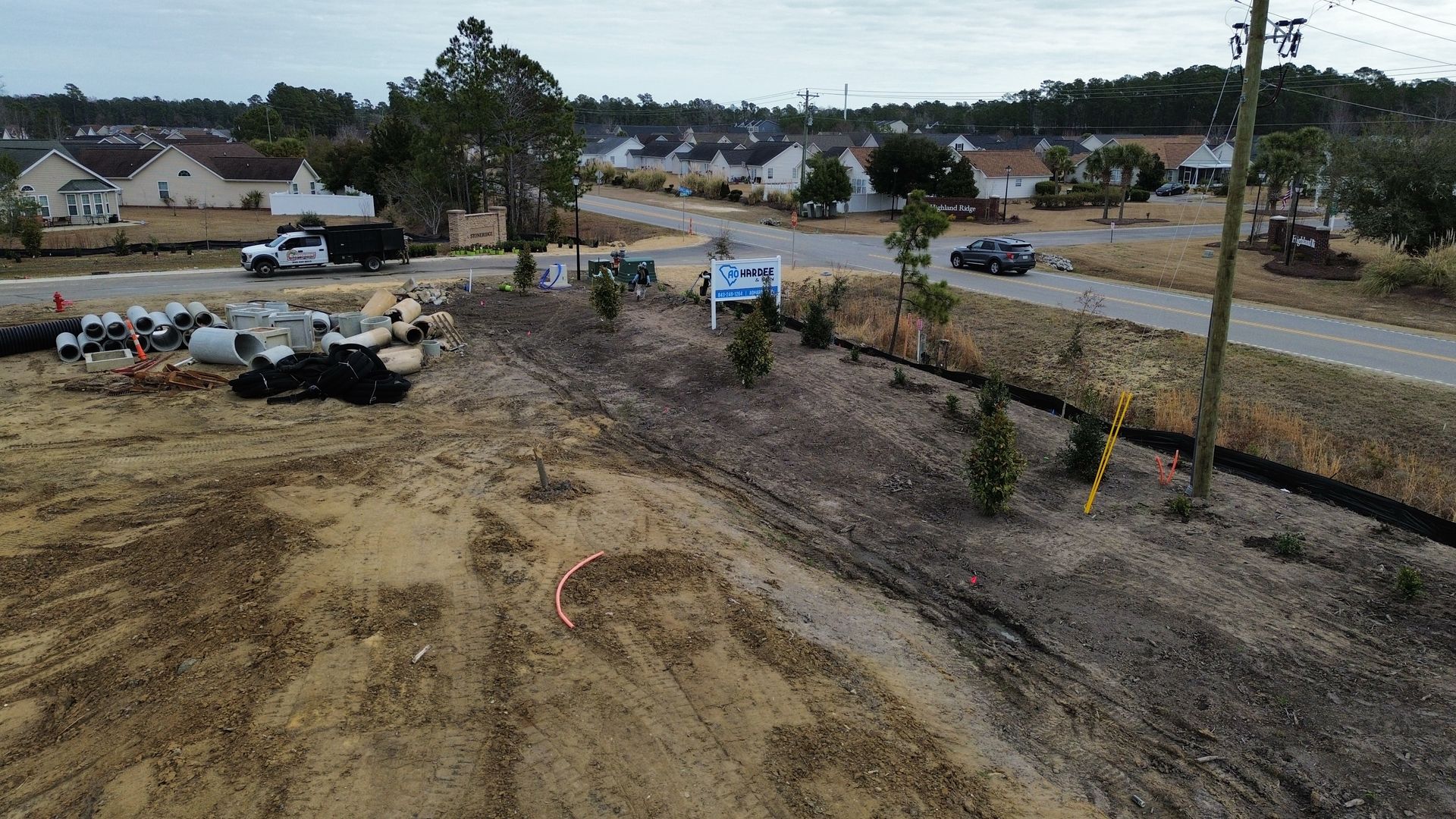 Construction site with dirt, trees, and houses in the background. A road and vehicles are visible.