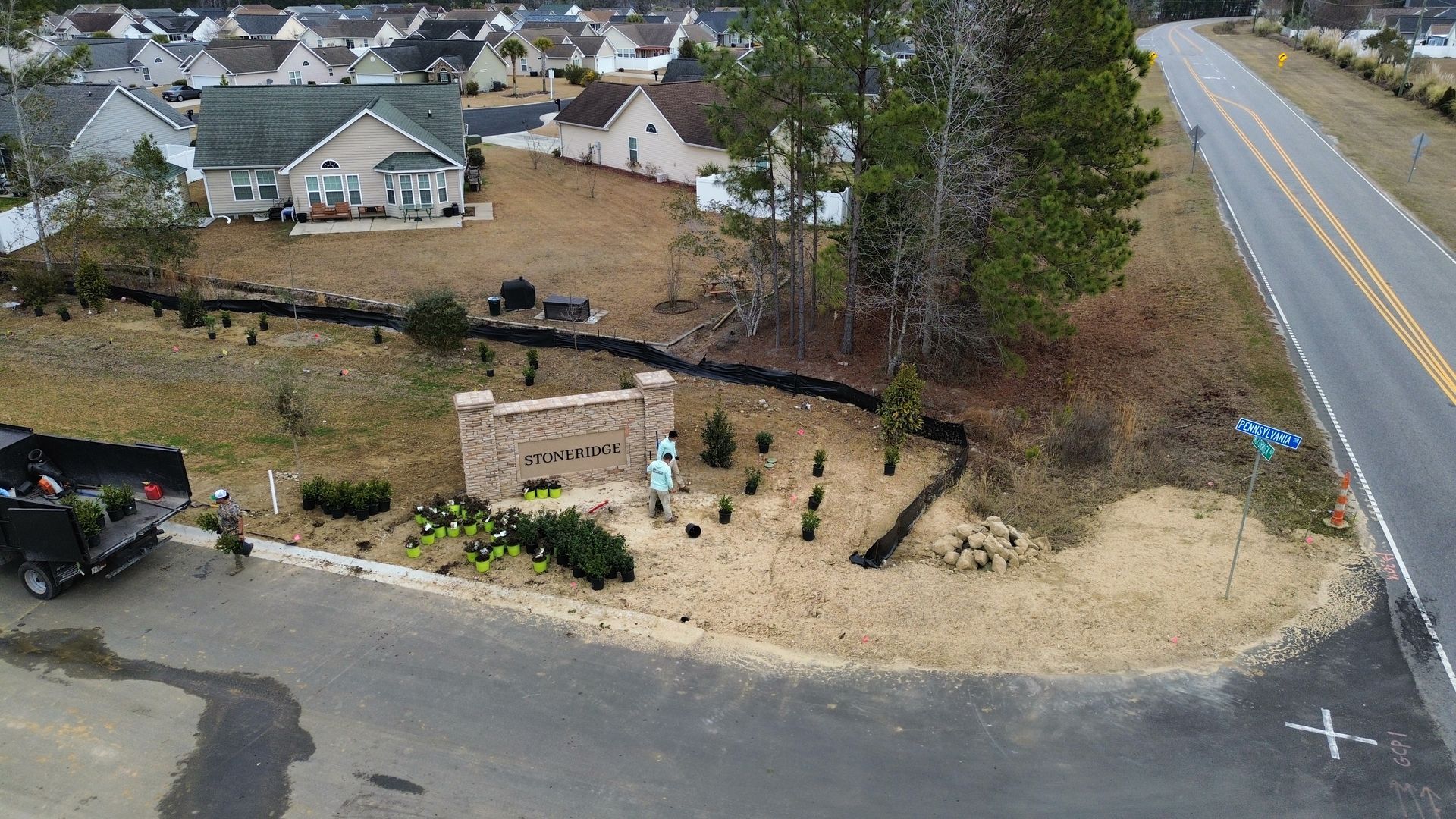 Construction workers near an entrance sign and planting shrubs next to a road, residential neighborhood in the background.