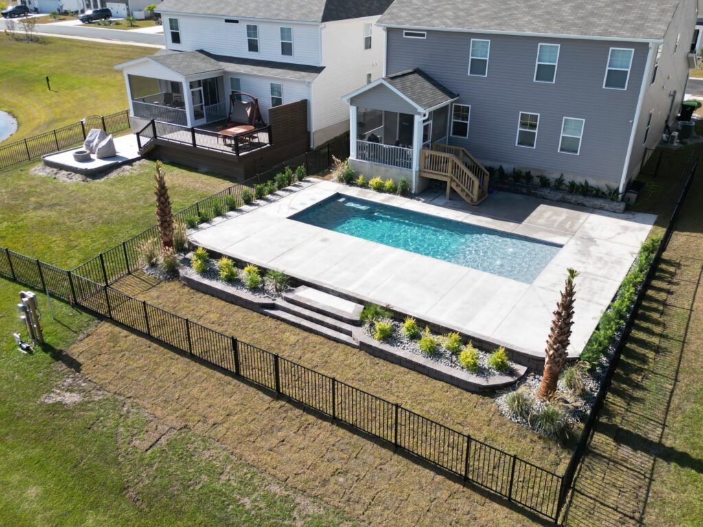 Backyard pool with black fence, two-story house, screened porch, landscaping, and palm trees.