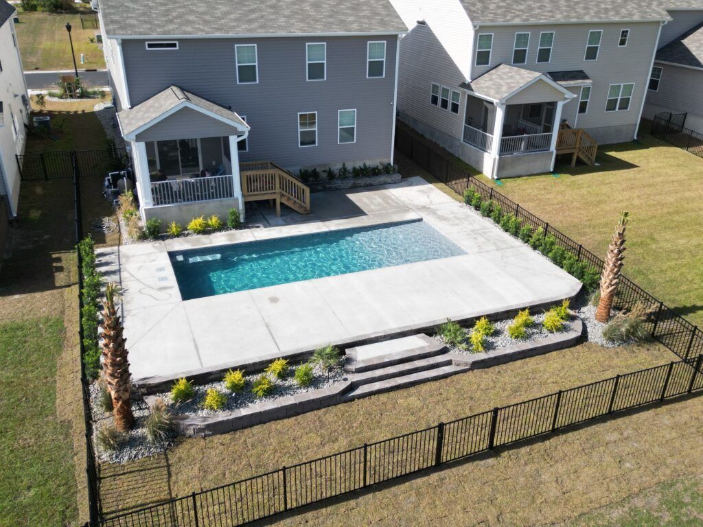 Backyard with a rectangular swimming pool, concrete patio, landscaping, and a gray house.