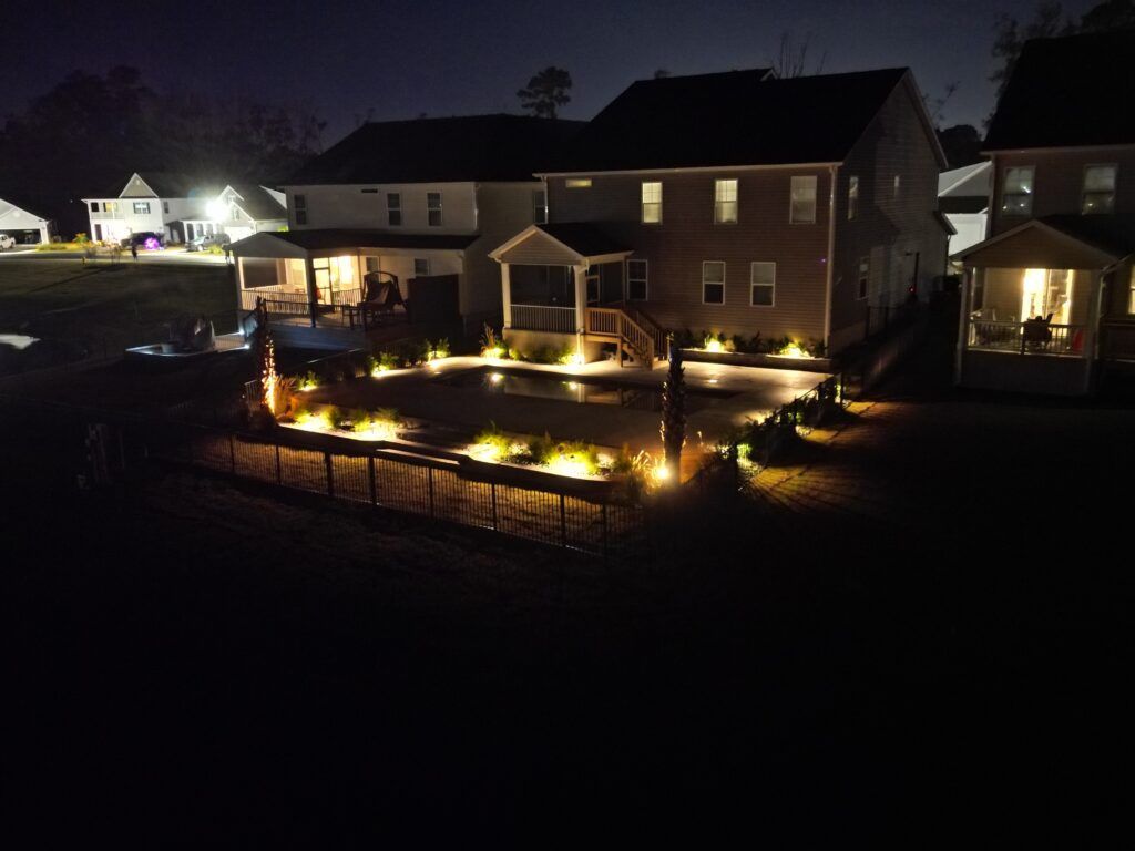 Backyard at night, illuminated by warm lights. Houses, landscaping, and a patio are visible.