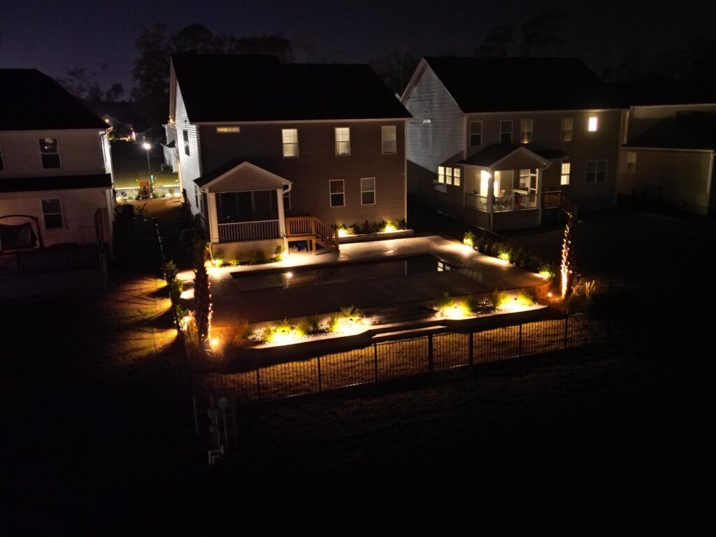 Night view of a backyard with a lit pool, landscaping lights, and houses.
