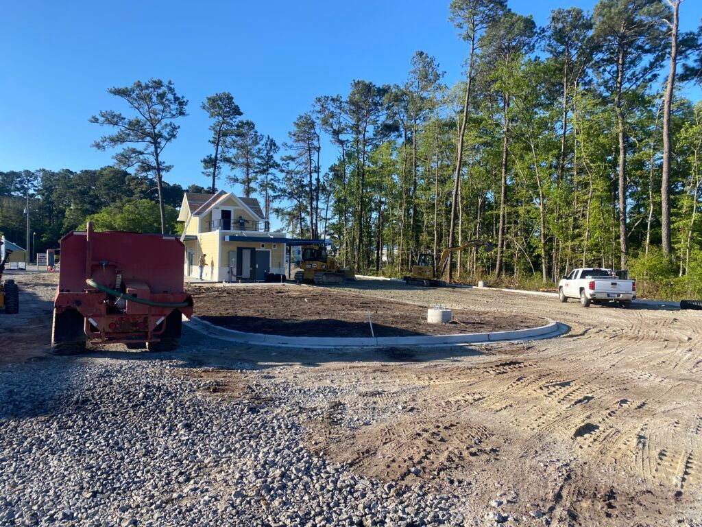 Construction site with a partially built house, gravel ground, and trees. A red machine and white truck are present.