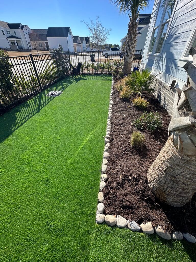 A narrow backyard with artificial turf lawn, a garden bed with mulch and stones, and a black fence.