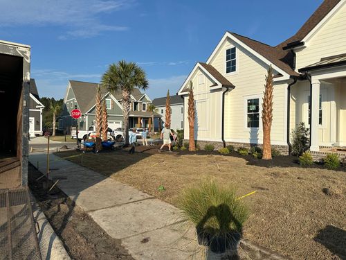 Workers install palm trees in front of a new, light-yellow residential home on a sunny day.
