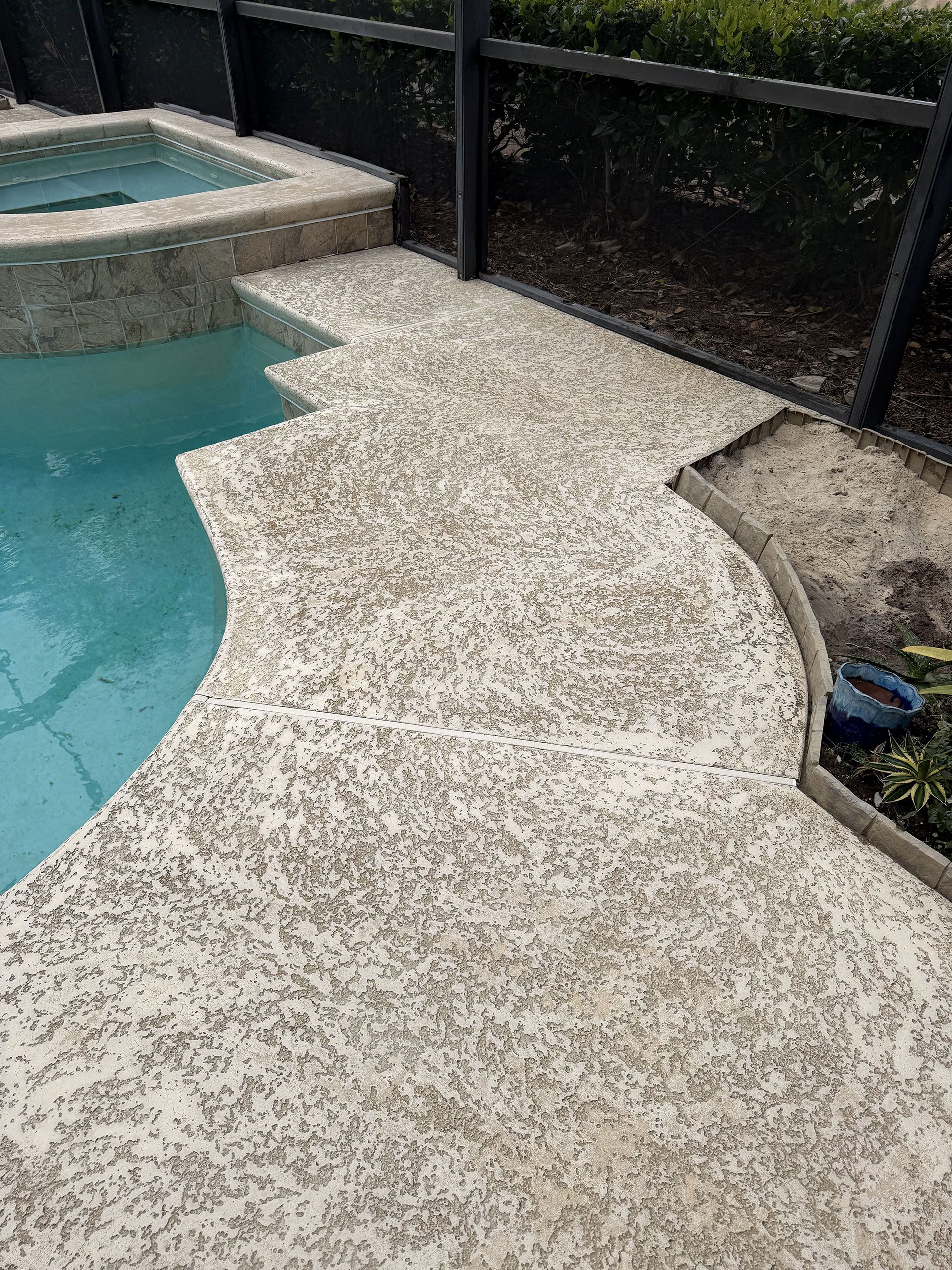 Curved white pool deck beside turquoise water and a black fence, with built-in steps and planter edge