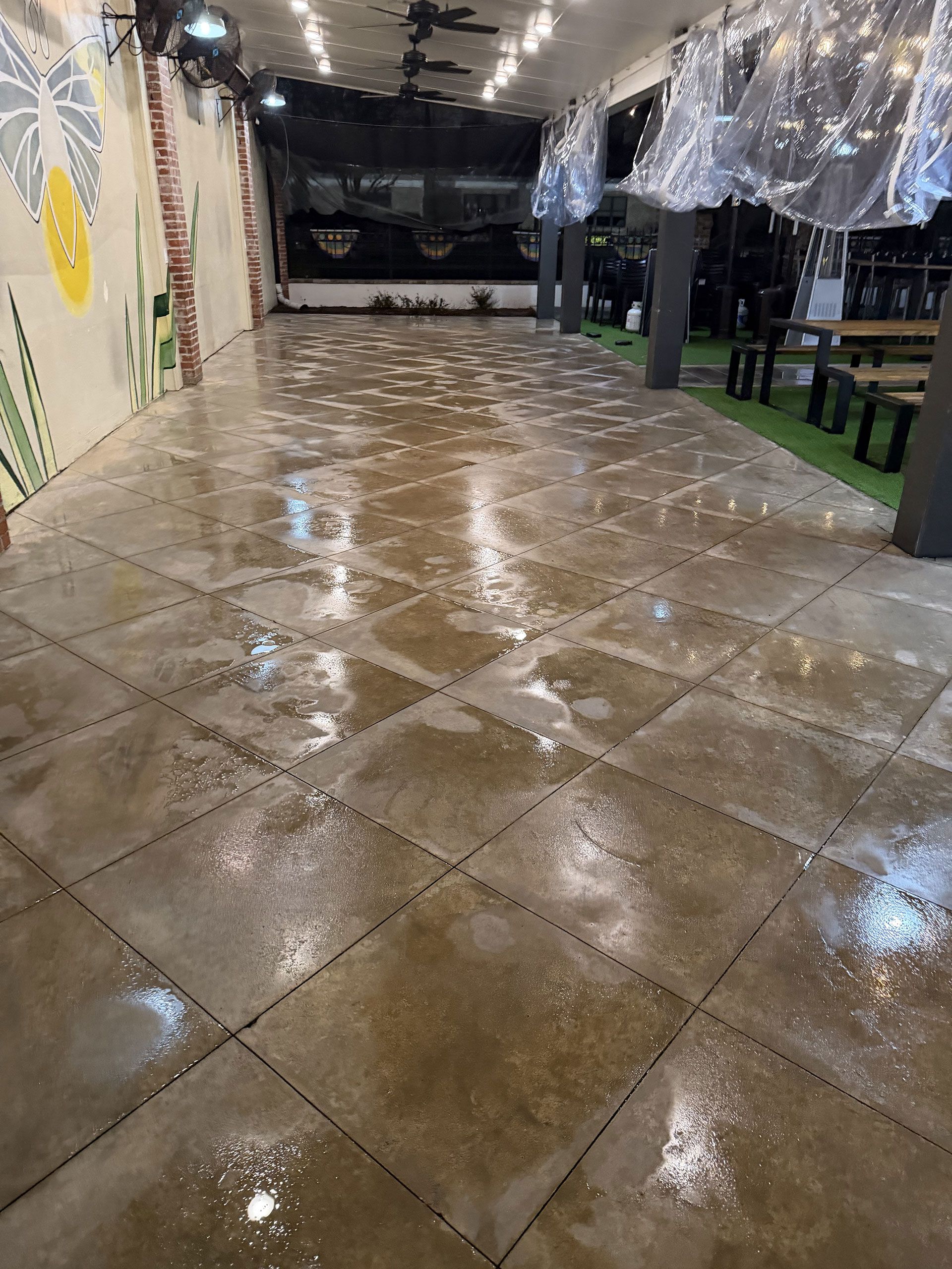 Wet tiled walkway at night under string lights, beside a mural wall and covered seating area.