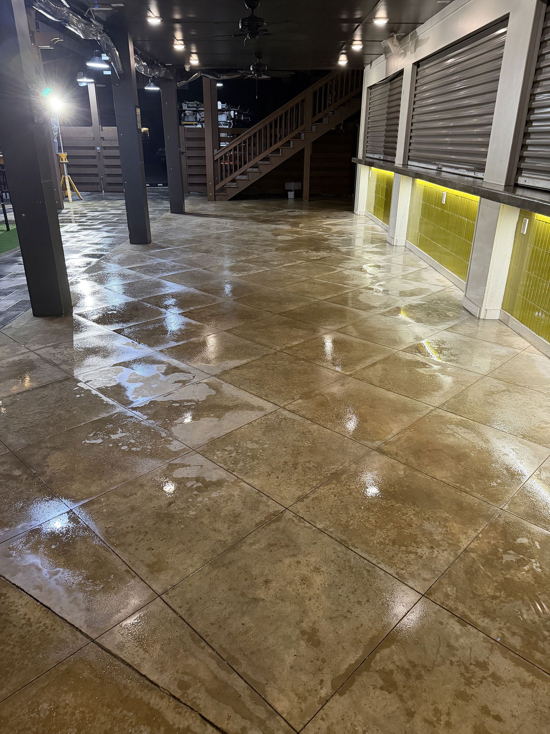 Flooded covered walkway with wet concrete, dark columns, and stairs under bright lights at night
