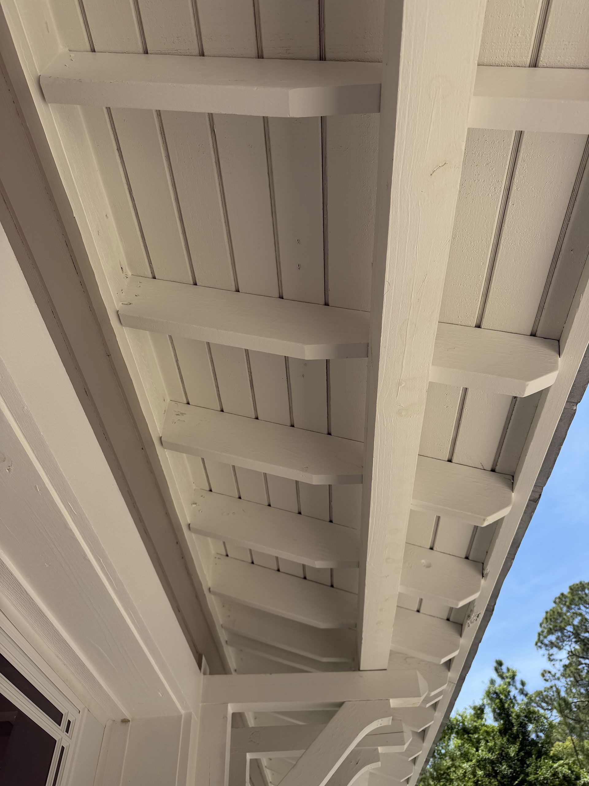 Close-up of white painted porch ceiling and support beams with blue sky and trees at the edge