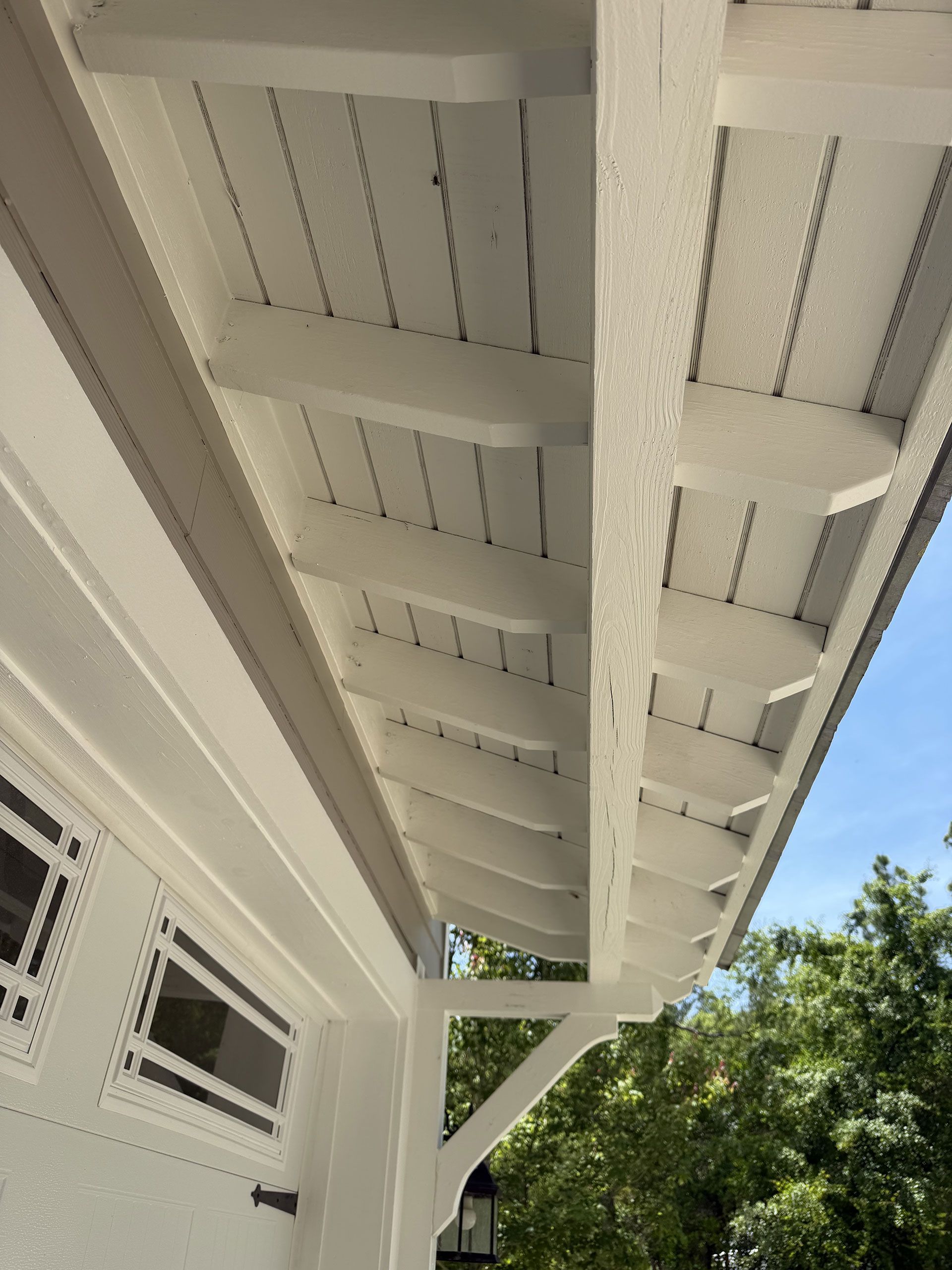 White house roof overhang with exposed rafters and a support brace against a blue sky and trees