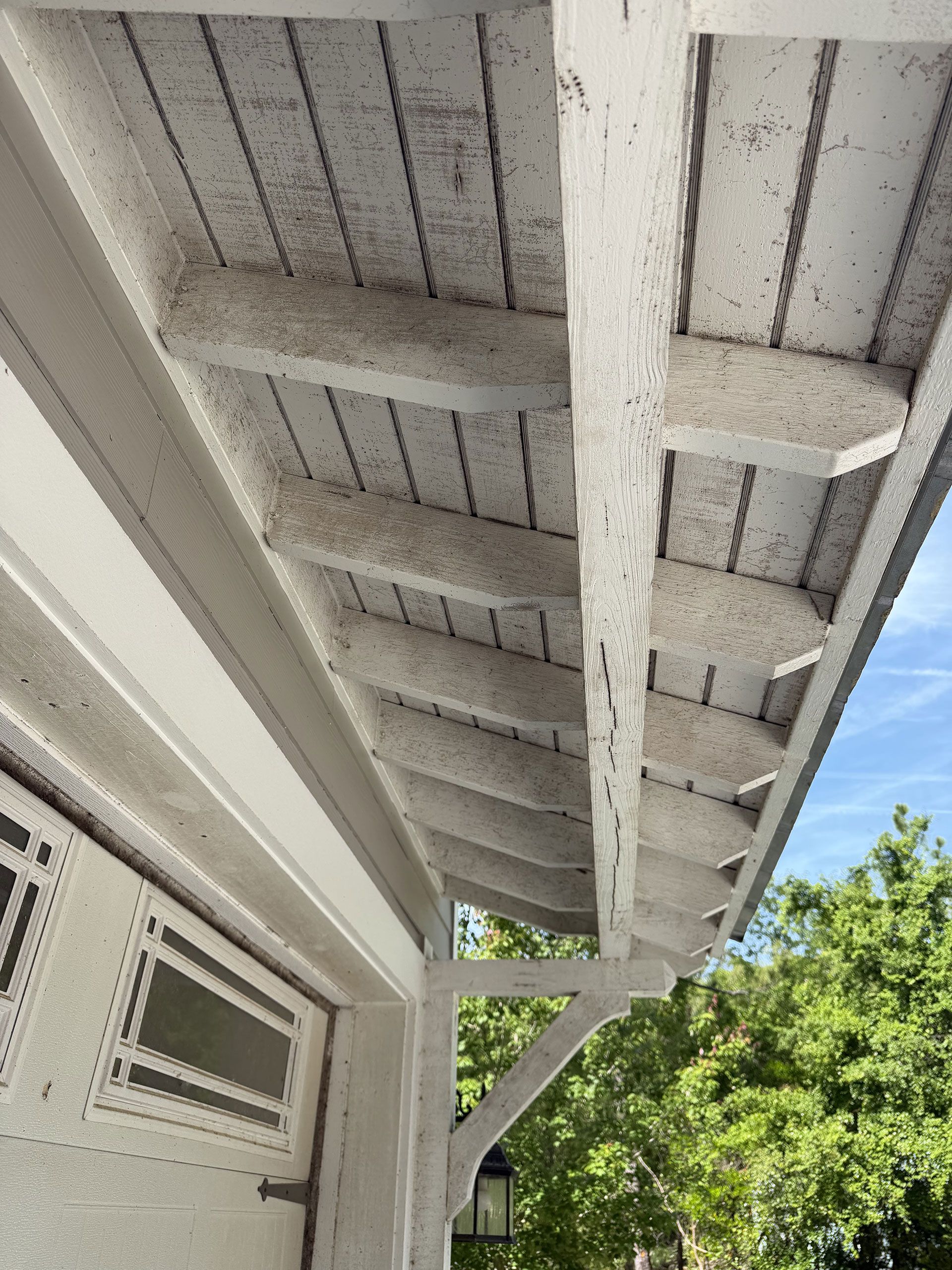 White wooden porch roof and support beams beside a house, with a window and green trees under a blue sky.