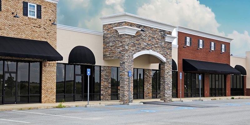 Row of empty storefronts with brick facades, arched entrances, and black awnings on a sunny street