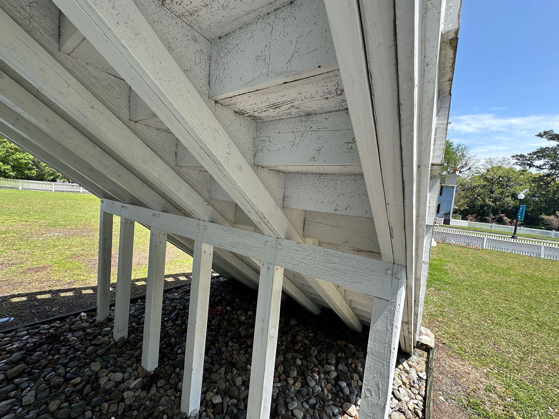 Underside of a white wooden staircase on posts, outdoors on gravel beside grass and blue sky.