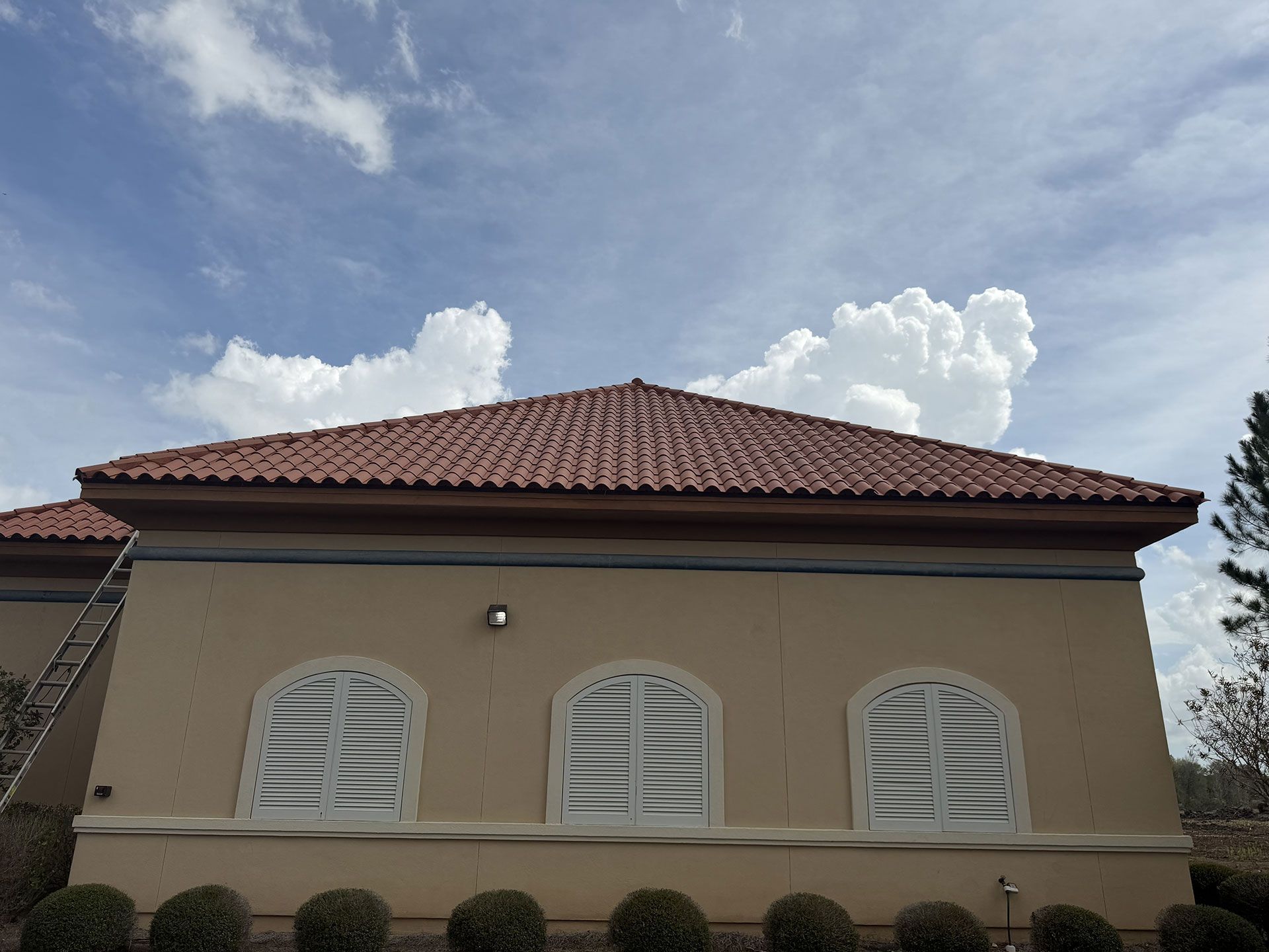 Beige house with red tile roof and white shuttered windows under a cloudy blue sky