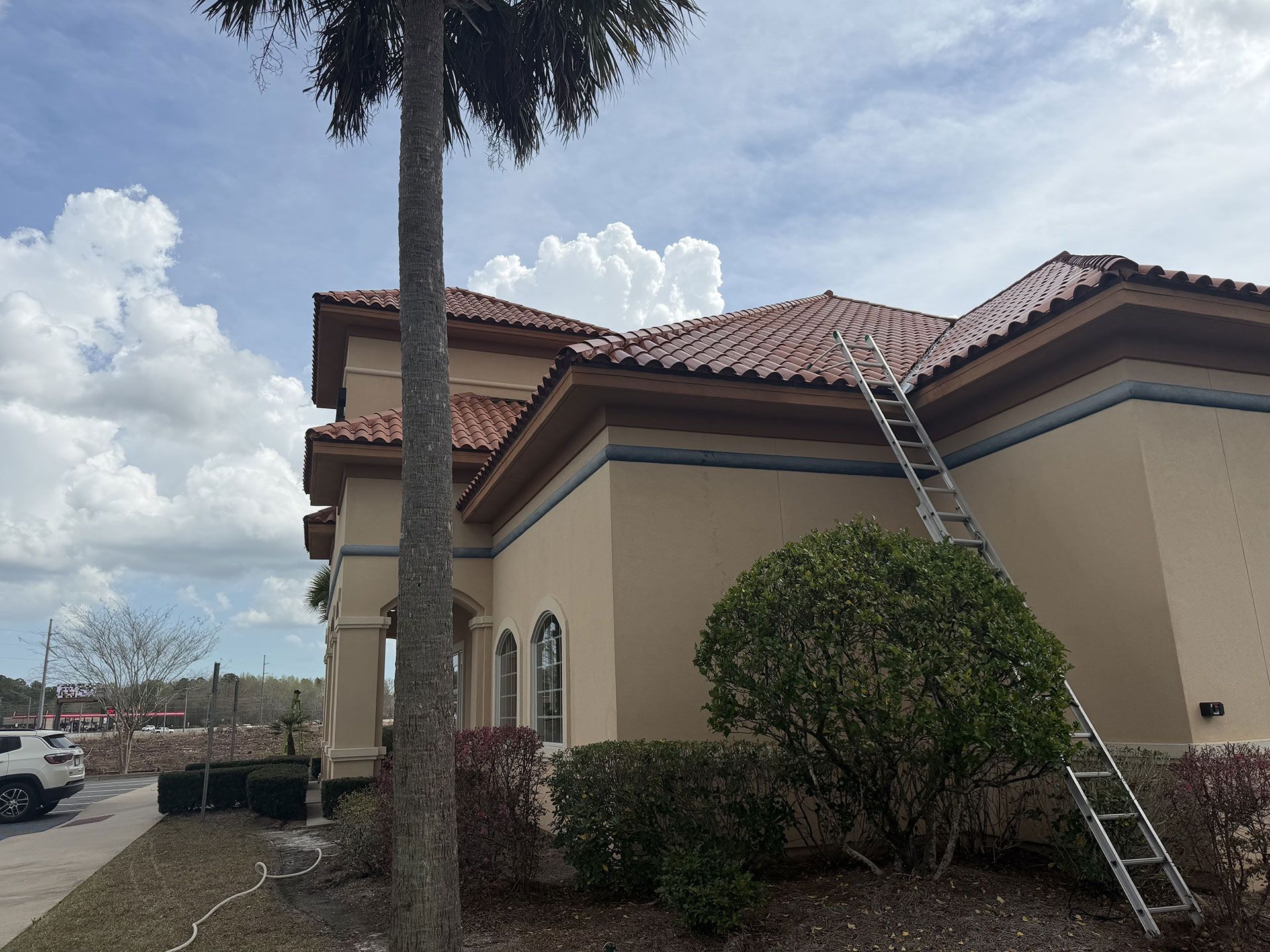 Single-story beige house with red-tiled roof, shrubs, and a ladder leaning against the wall under a cloudy sky