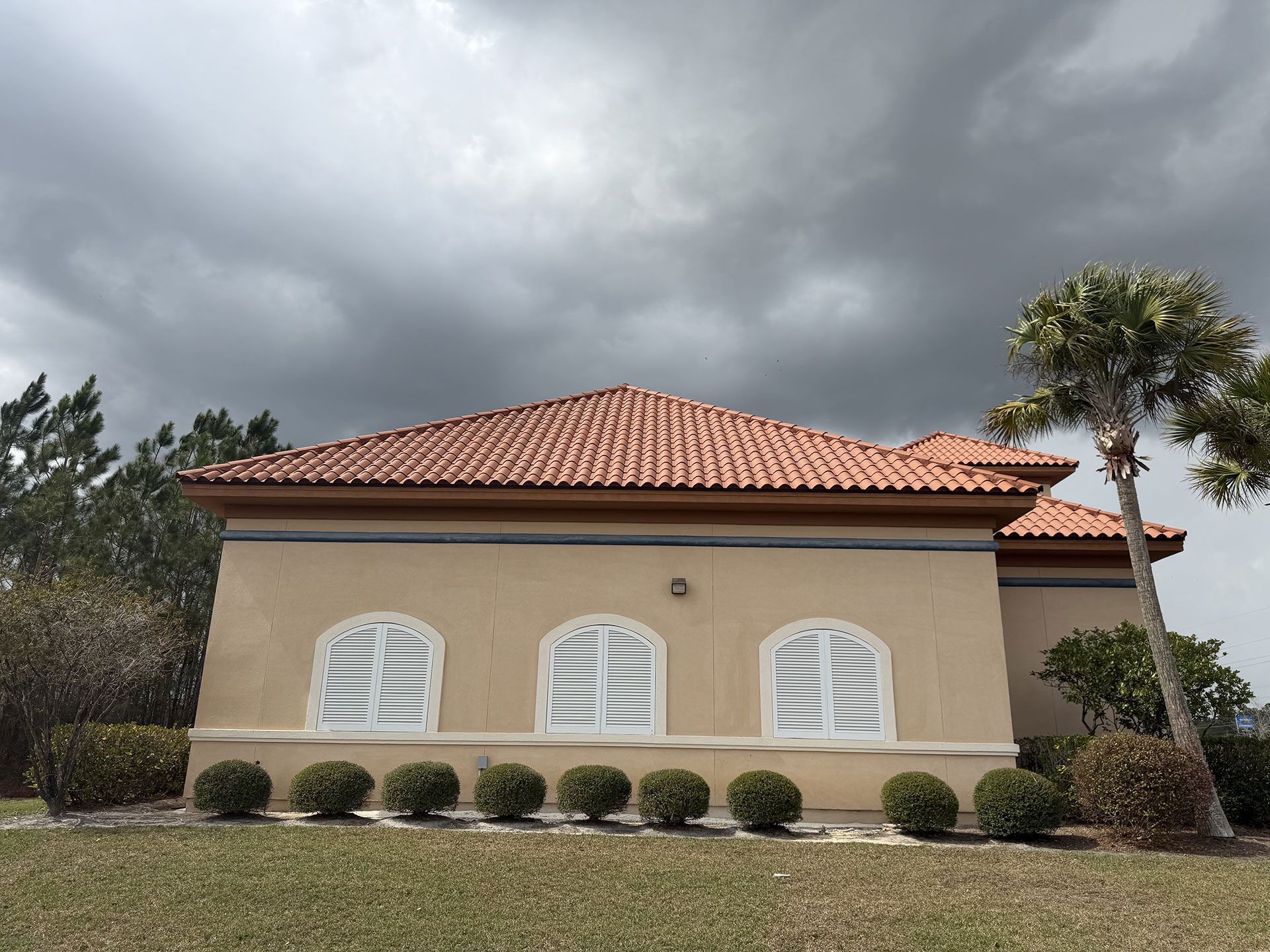 Single-story beige house with terracotta roof and white shutters under dark storm clouds