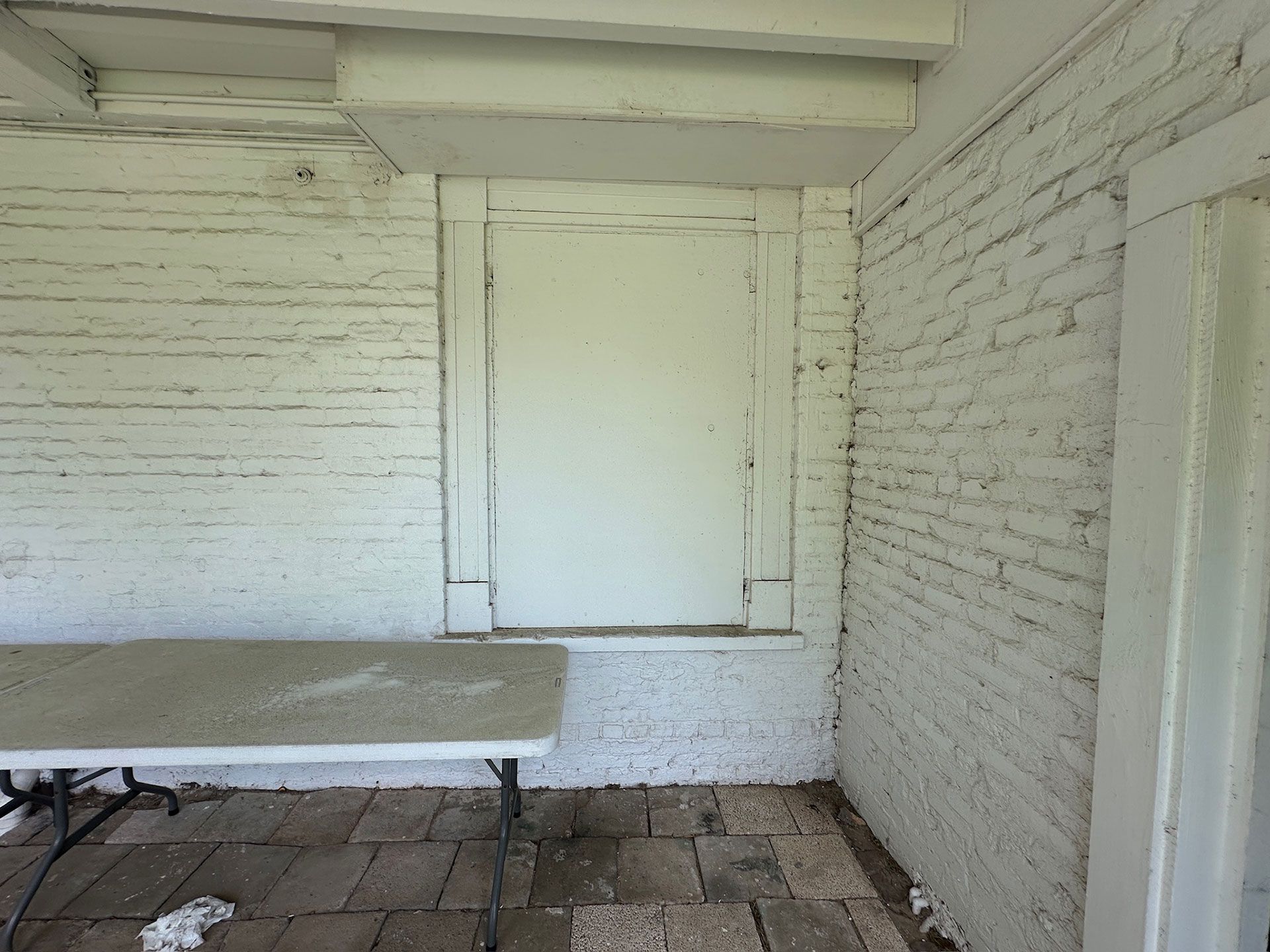Small covered porch with white brick walls, a closed door, and a folding table in the foreground.