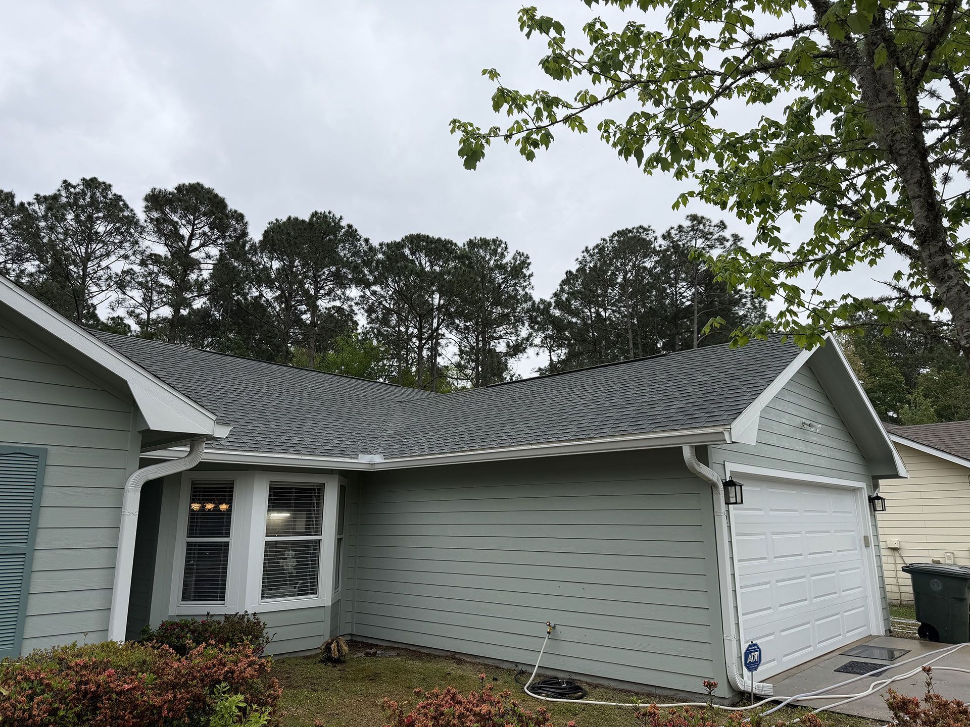 Single-story house with damaged, wavy roof shingles and attached garage in a wooded yard