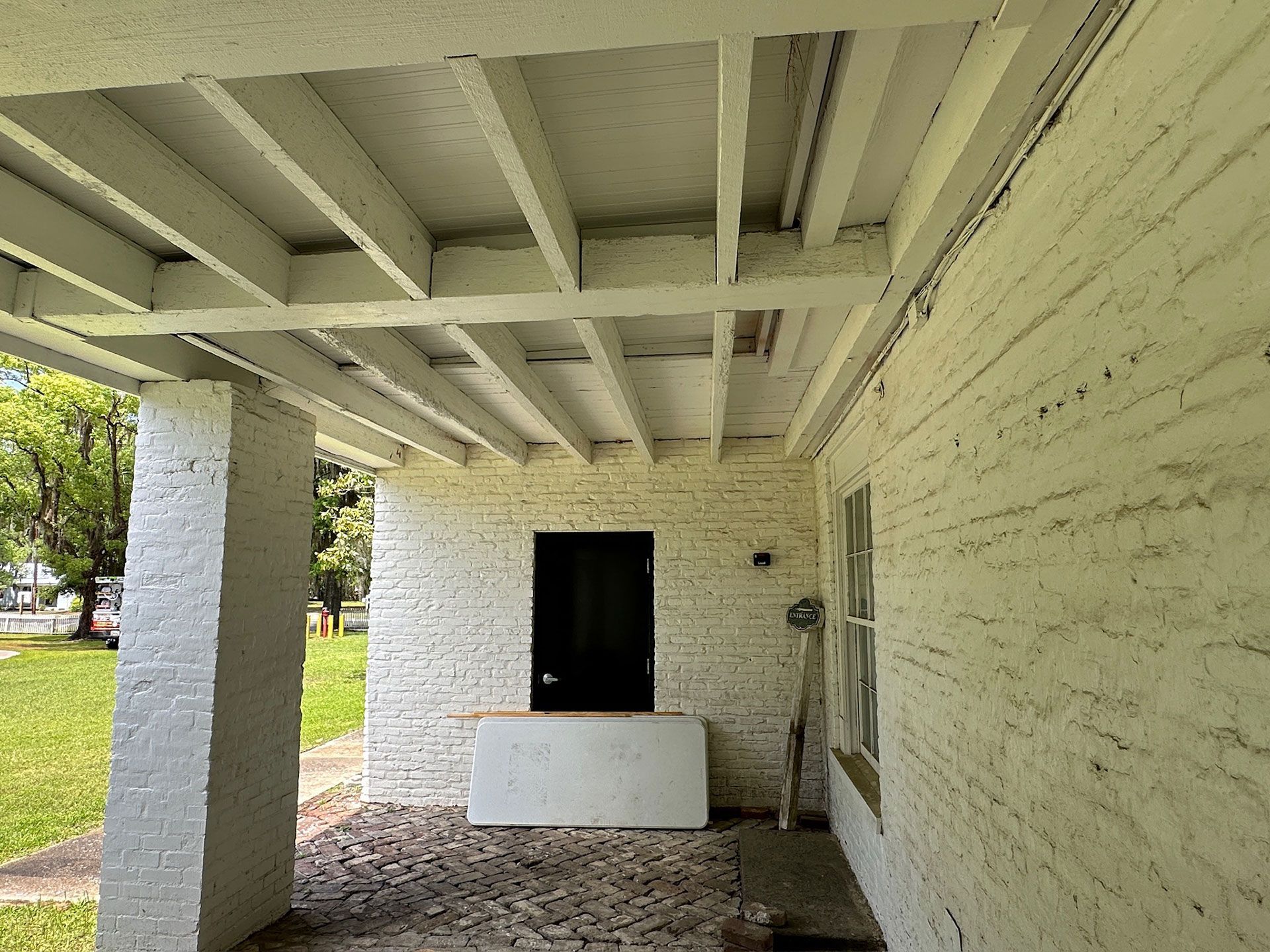 Covered porch with white-painted beams, brick wall, black door, and a white bench in the shade.