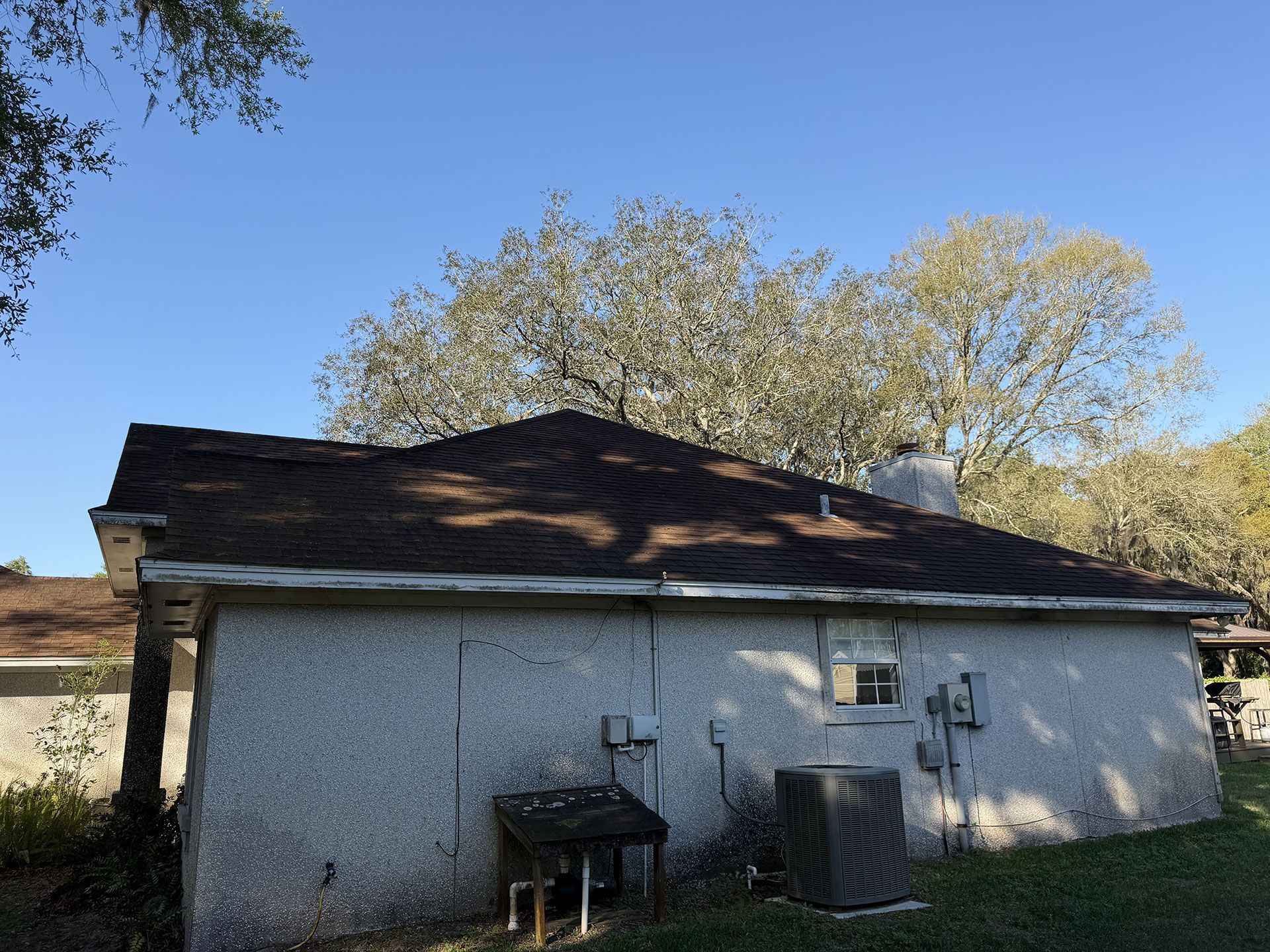 Small gray house with dark roof, air conditioner, and grill in a backyard under a blue sky.