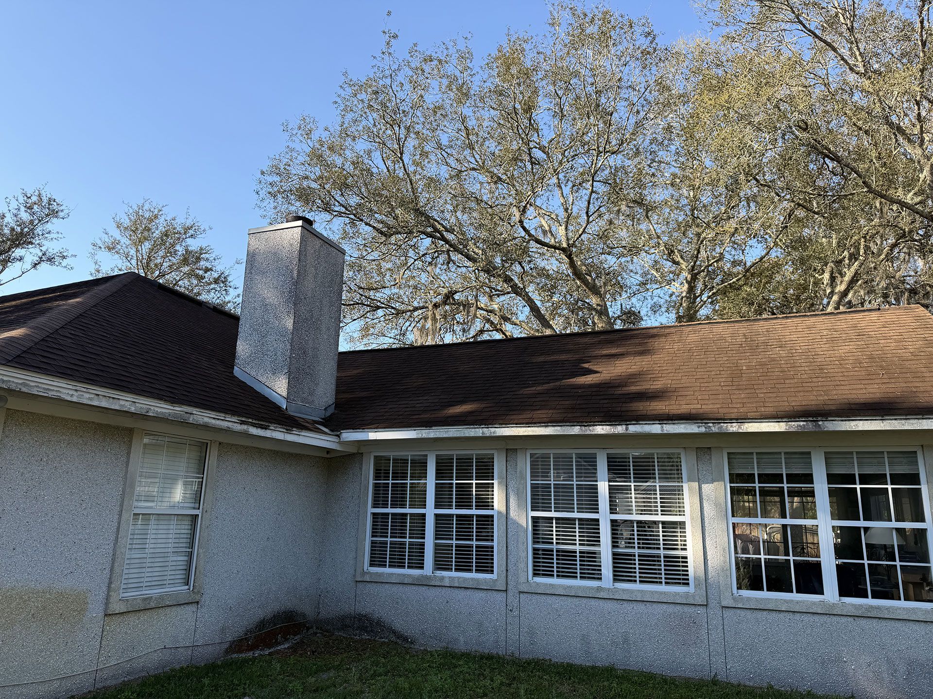Single-story stucco house exterior with white windows, brown roof, chimney, and a tree behind it