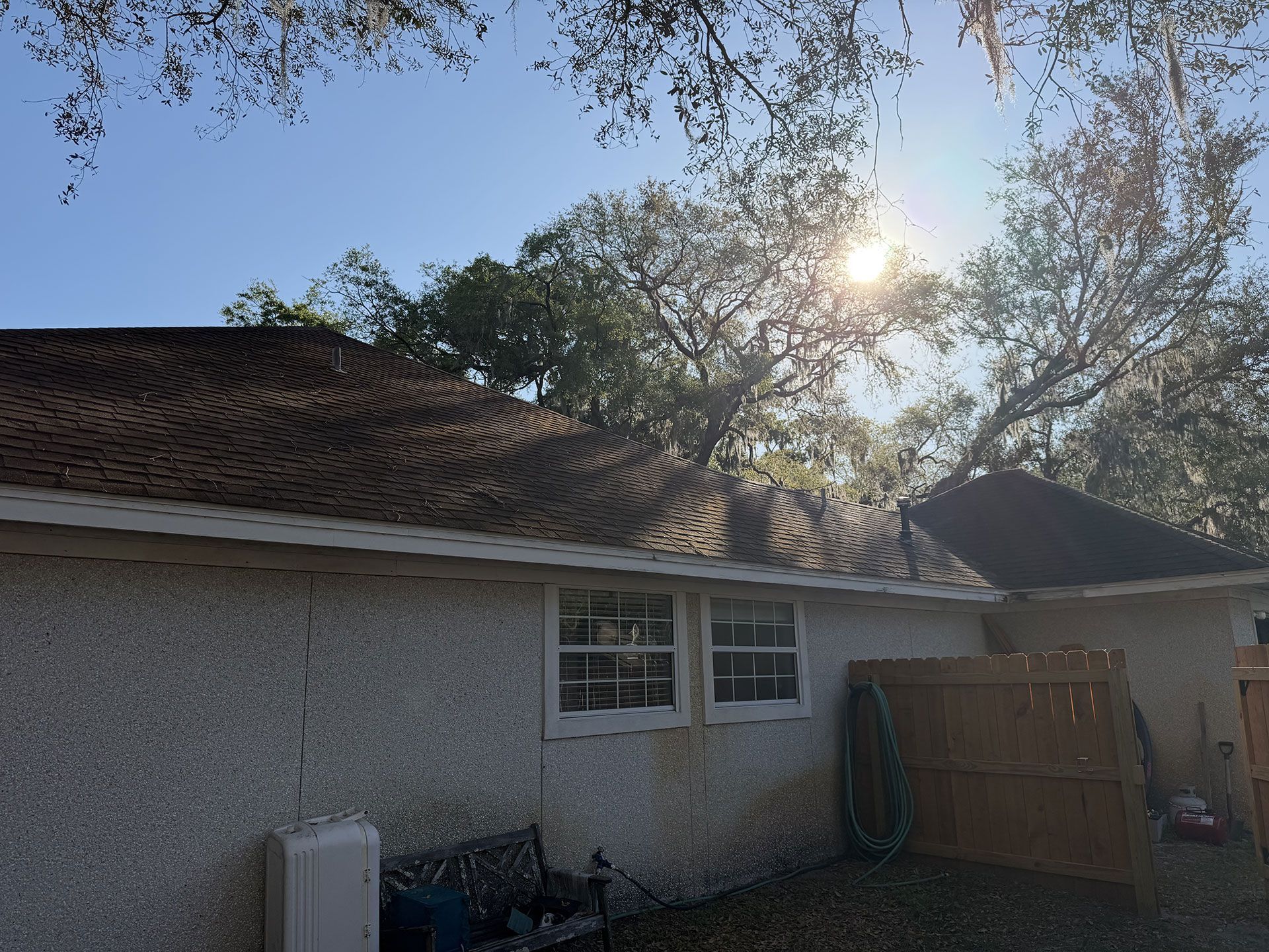 Sunlit house exterior with stucco walls, tiled roof, trees overhead, and a wooden gate