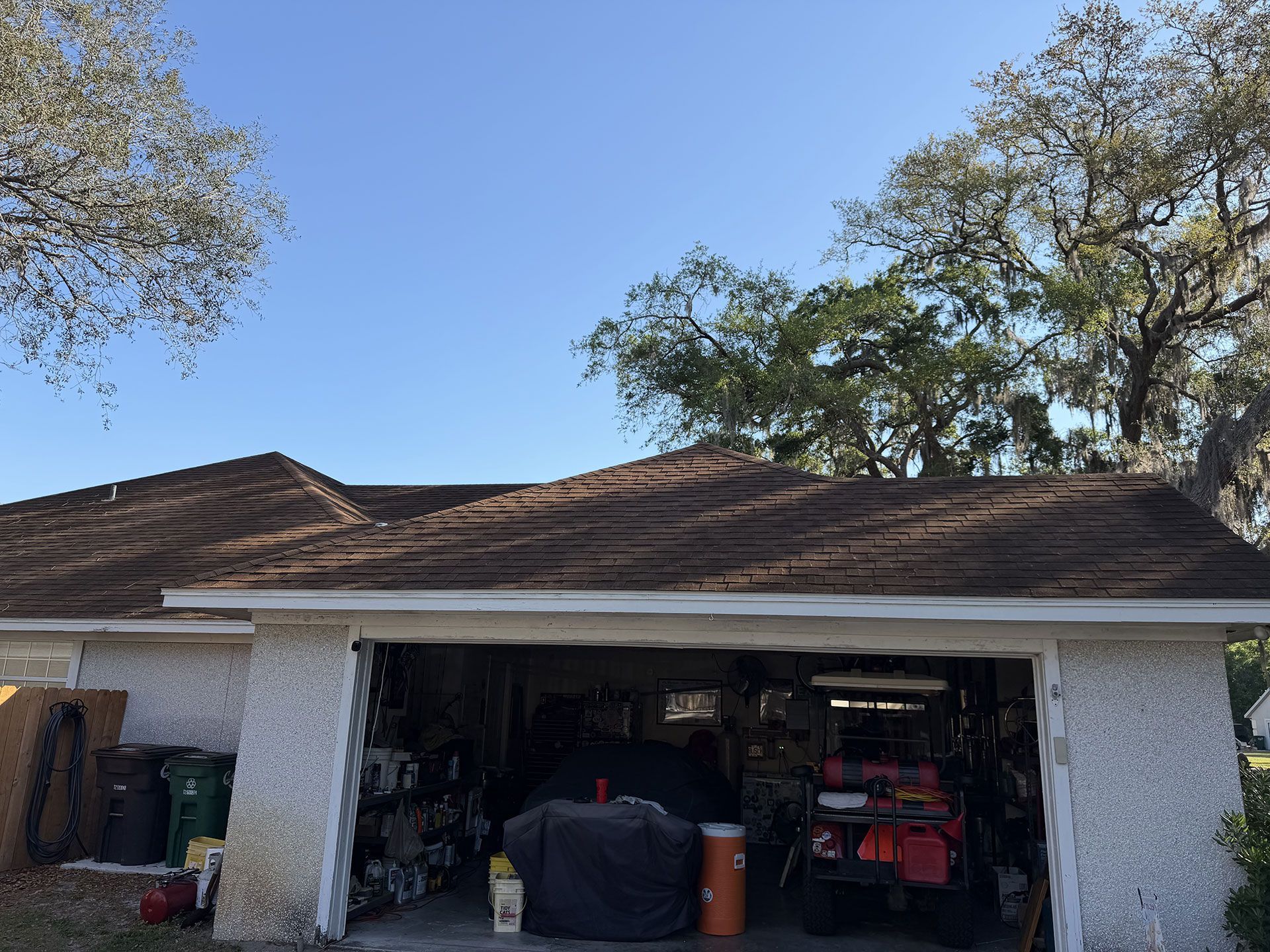 Open garage with tools and covered items under a brown shingled roof, framed by trees and blue sky