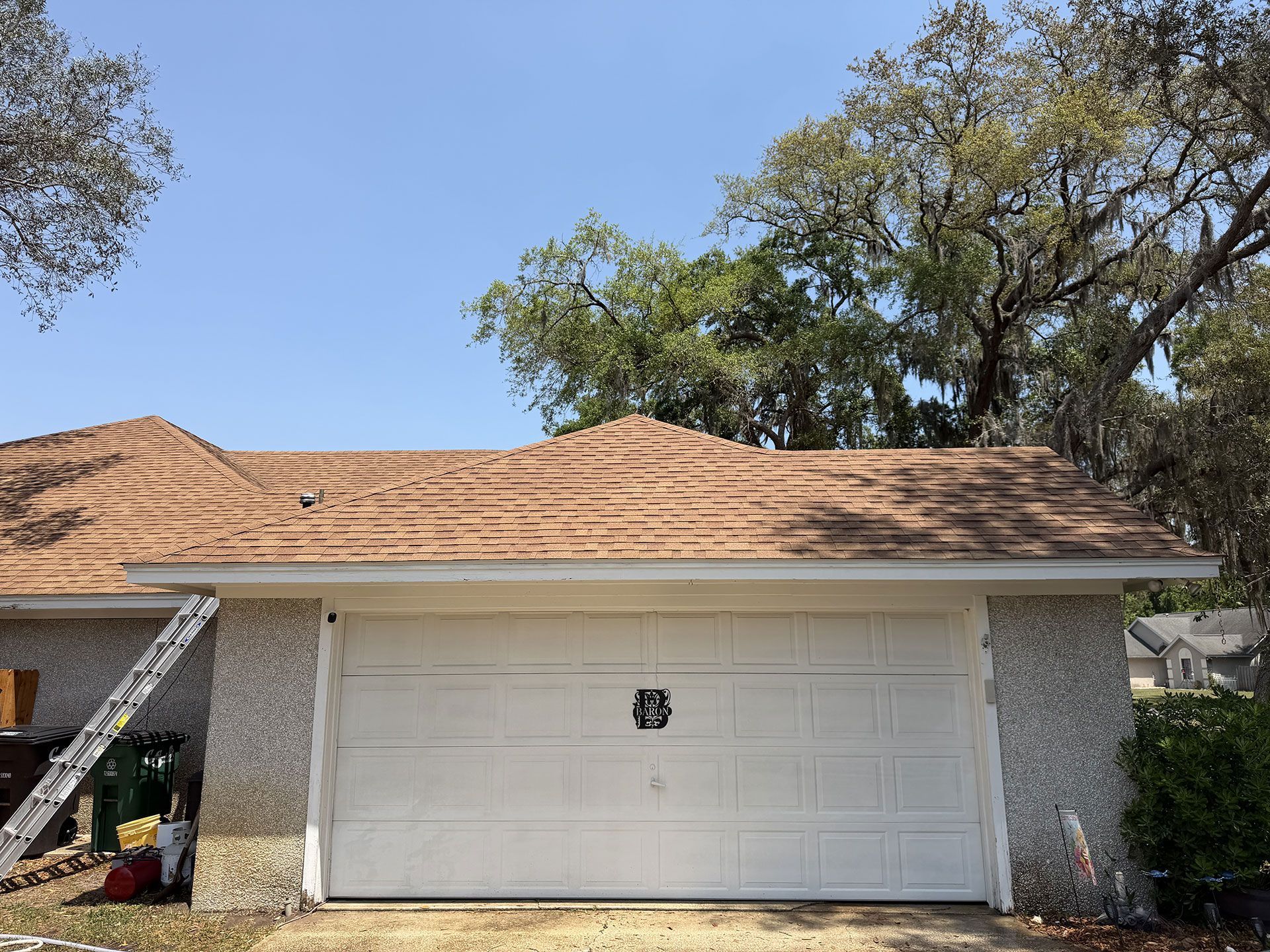 Single-story house exterior with tan roof, white garage door, and trees under a clear blue sky
