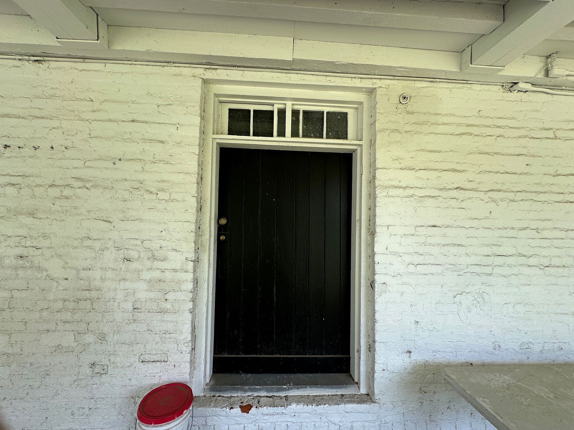 Black front door in a white brick wall with a small red stool on the porch.