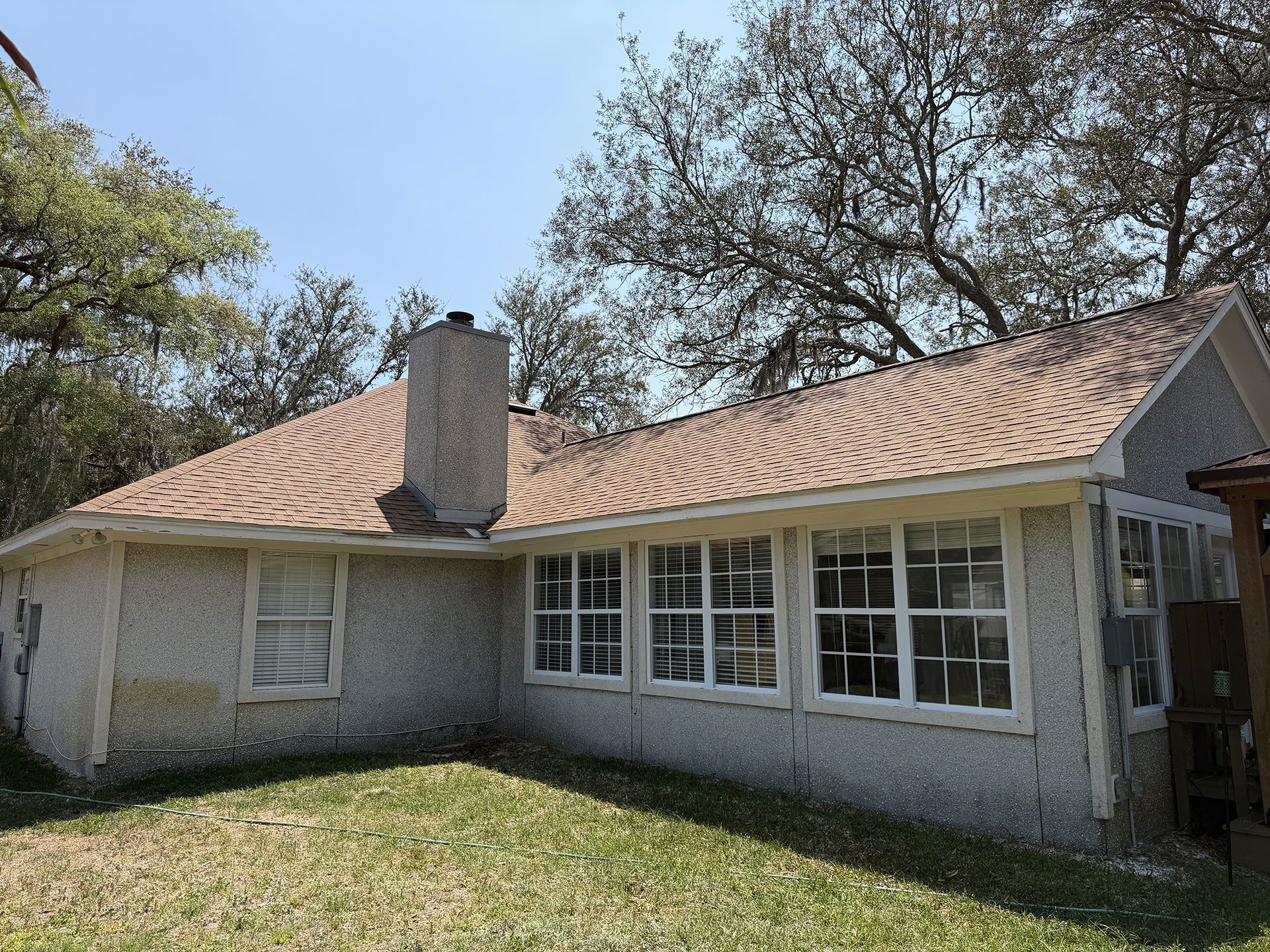 Single-story brick house with tan roof, chimney, and large front windows under trees