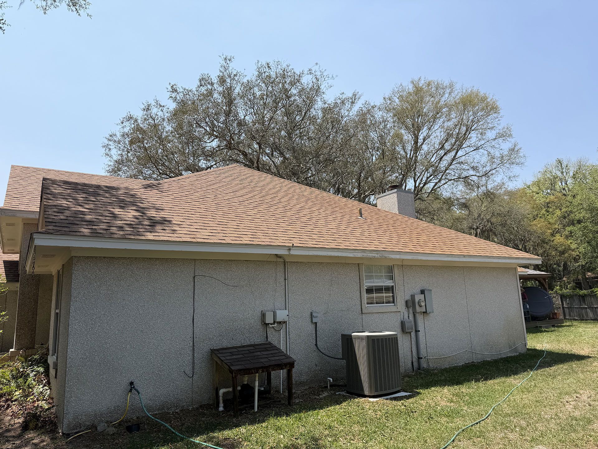 Single-story house exterior with tan shingle roof, gray stucco walls, and air conditioner units in a grassy yard
