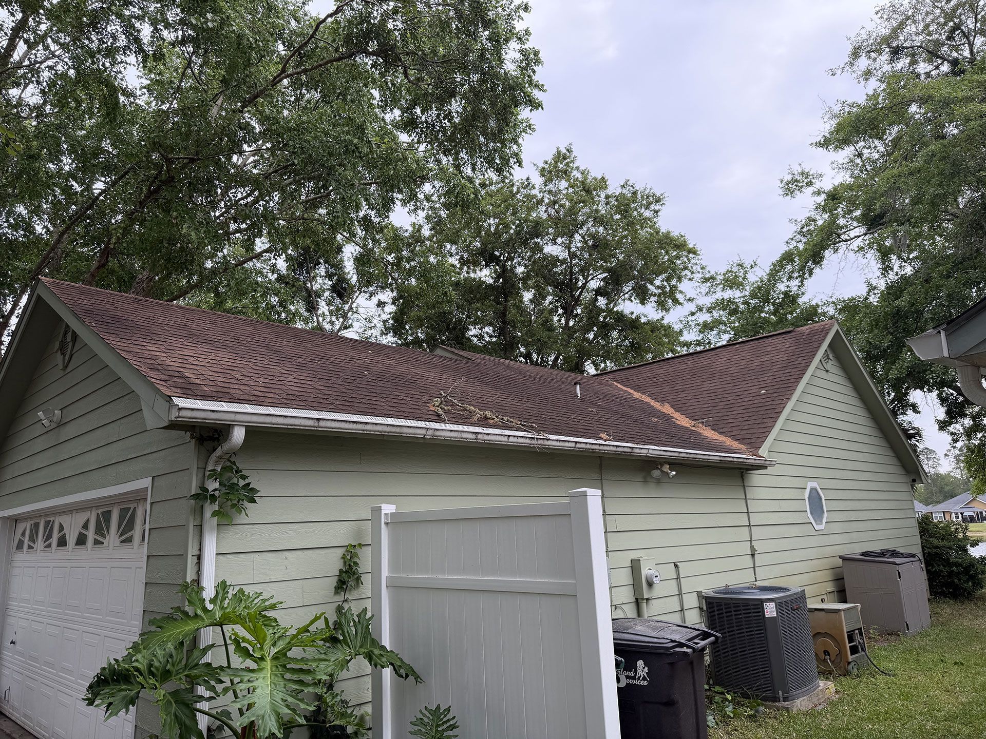 White garage with brown shingle roof and small shed, surrounded by trees and cloudy sky.
