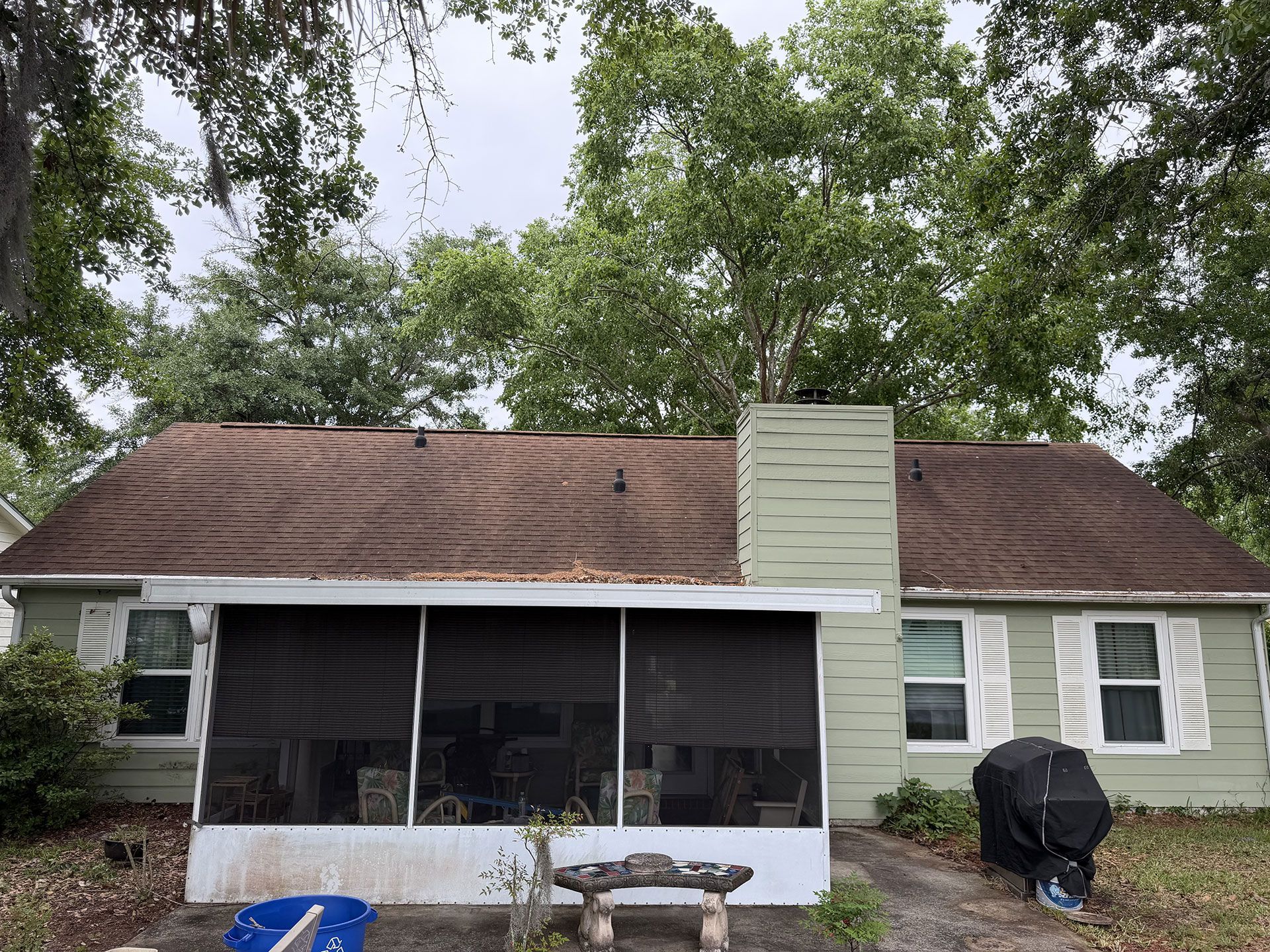 Small beige house with a brown roof, screened porch, and a grill in the yard under tall trees.