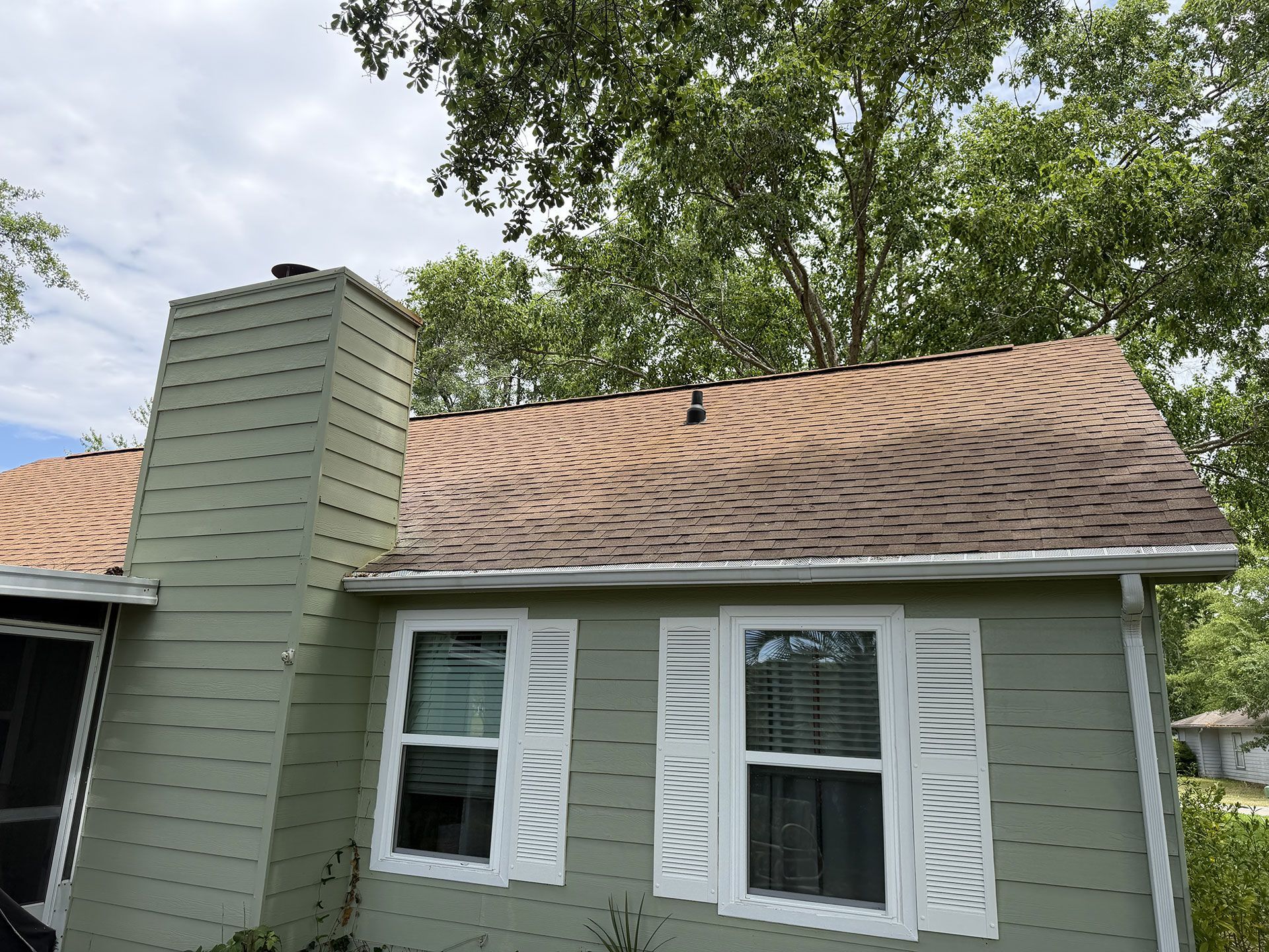 Small gray house with tan shingle roof, white-trim windows, and a chimney under leafy trees.