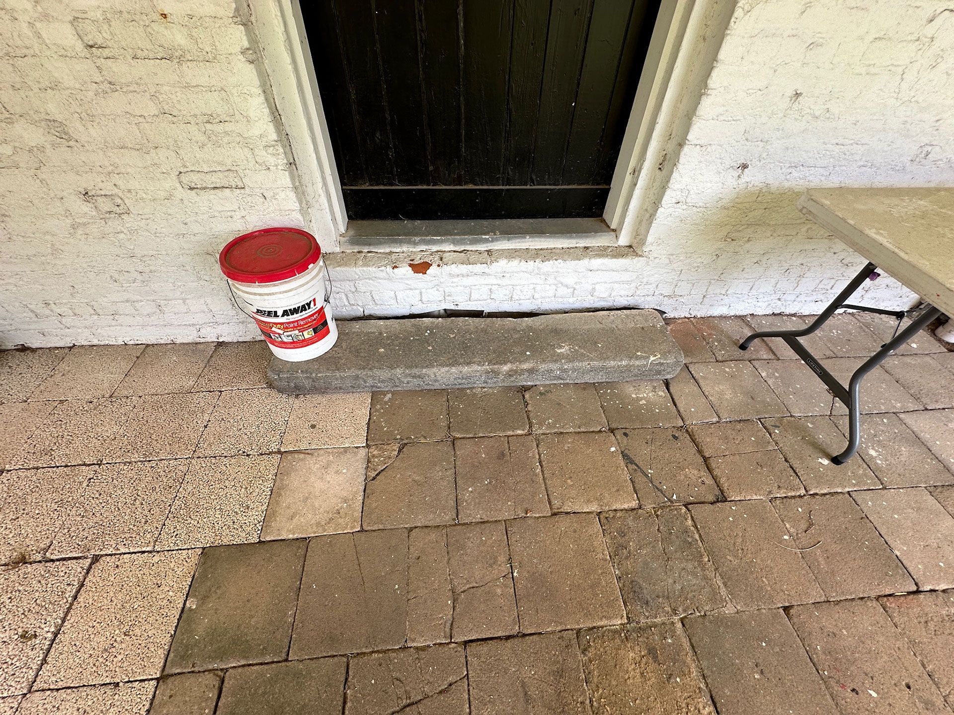 Porch entrance with red bucket beside a doorway and a chair on brick pavers