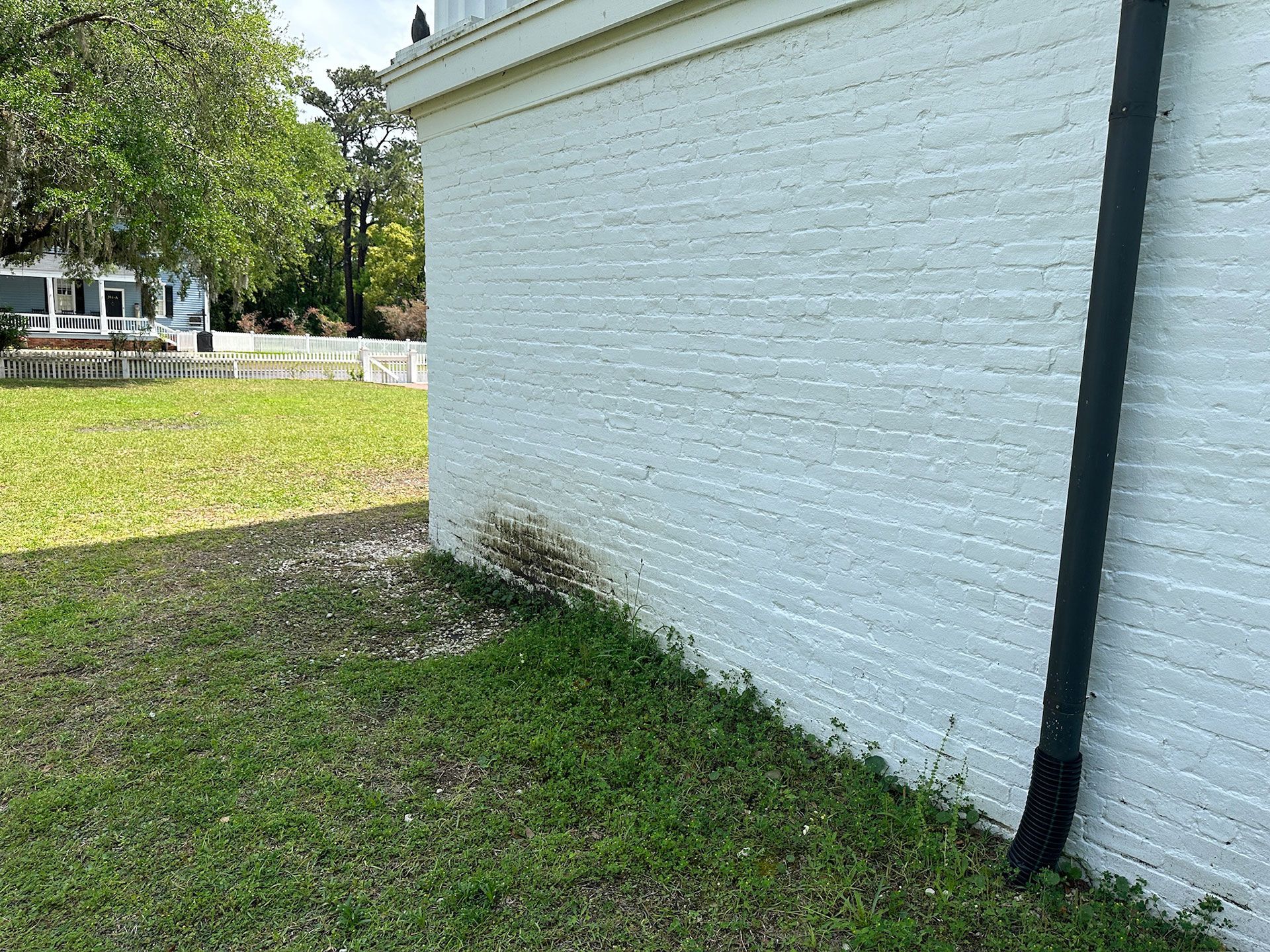 White brick wall corner with a black downspout beside a grassy yard and trees in the background